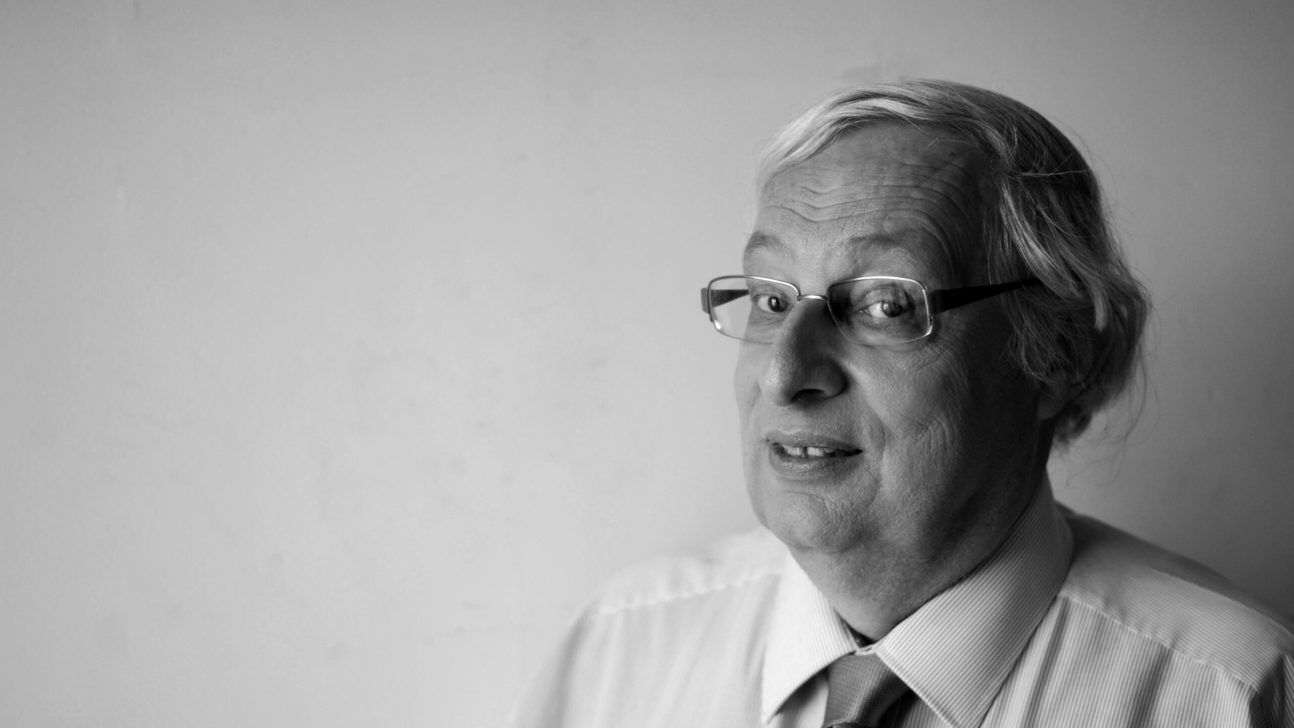 L'image montre un homme d'âge moyen avec des cheveux gris et des lunettes. Il porte une chemise claire avec une cravate. Il regarde légèrement sur le côté avec un sourire, le fond étant neutre et clair. L'image est en noir et blanc, ce qui lui donne un aspect classique et intemporel.