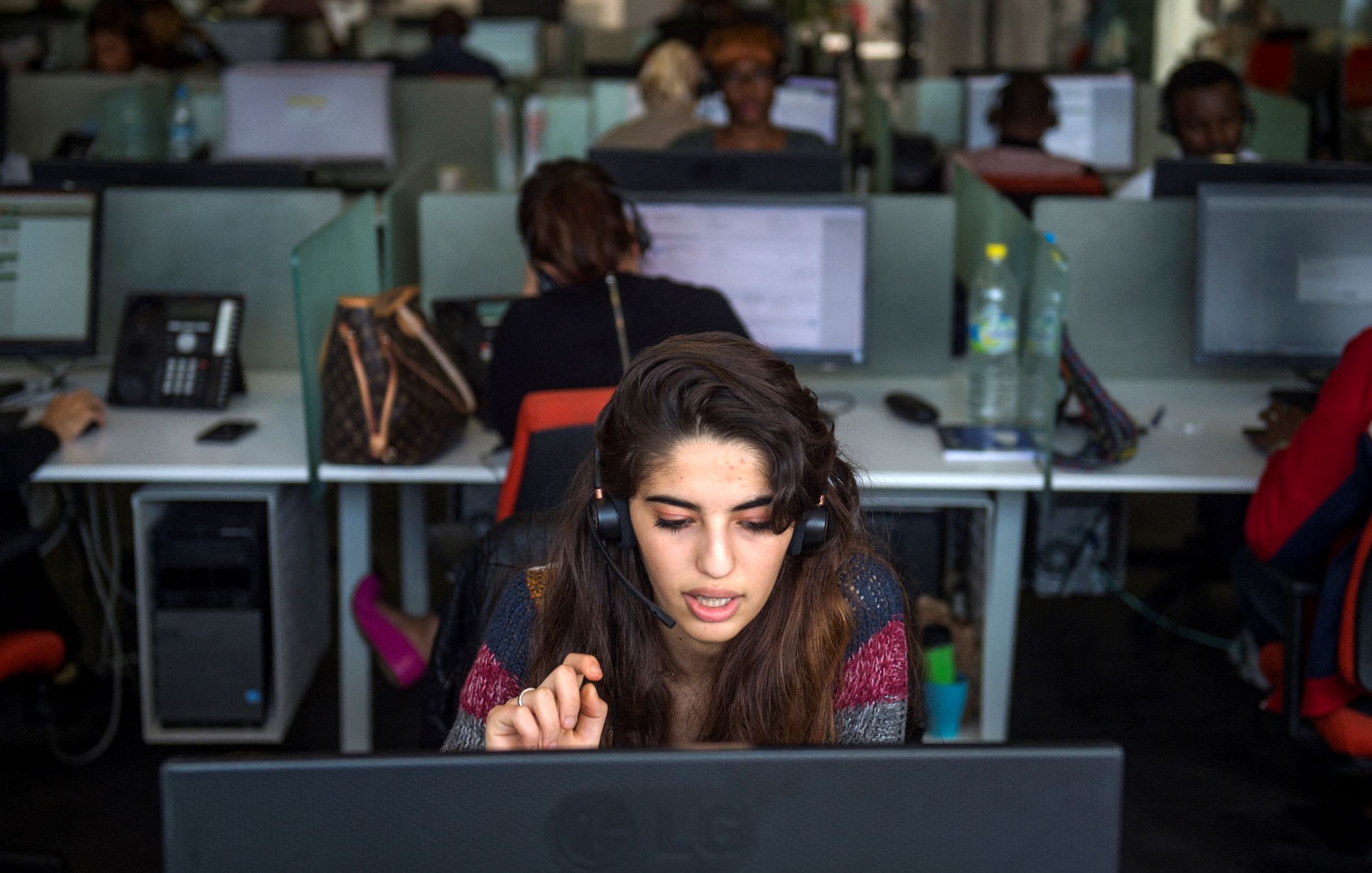 L'image montre un bureau avec plusieurs employés travaillant sur des ordinateurs. Au premier plan, une jeune femme porte un casque et semble concentrée sur son écran. Elle gesticule avec une main, probablement en train de communiquer avec un client. En arrière-plan, d'autres personnes sont visibles, toutes occupées à leurs tâches. L'environnement a l'air moderne, avec des bureaux en open space et des équipements de travail.
