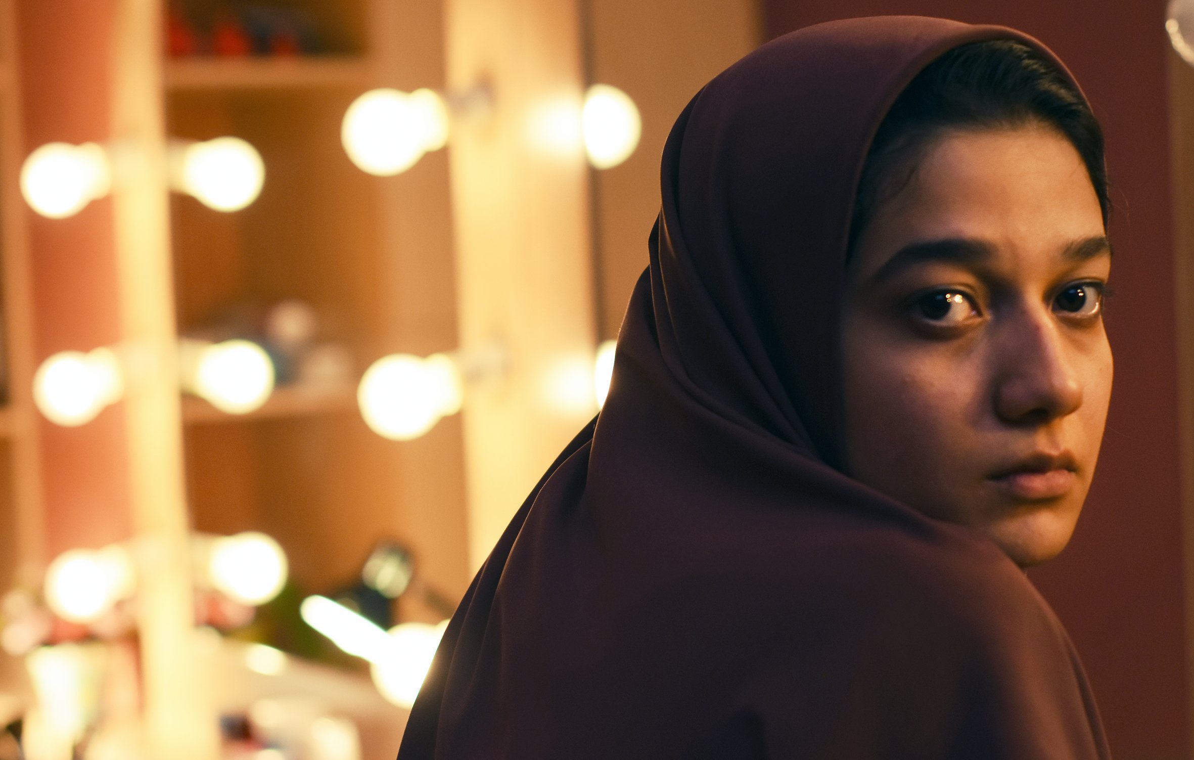 L'image montre une jeune femme avec un regard pensif, portant un hijab. Elle est positionnée de manière à ce que son visage soit tourné légèrement vers la caméra, tandis qu'en arrière-plan, on aperçoit un miroir entouré de lumières douces, créant une ambiance chaleureuse et intime. Son expression reflète une certaine introspection, ajoutant une profondeur émotionnelle à la scène.