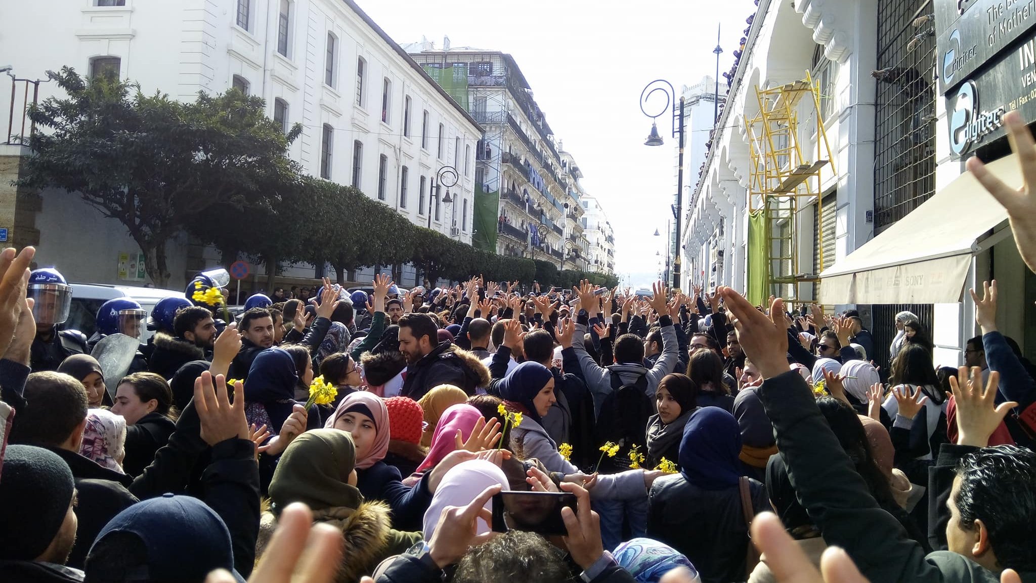 L'image montre une grande foule de personnes rassemblées dans une rue, levant les mains en signe de protestation ou de solidarité. On peut voir des bâtiments architecturaux en arrière-plan, ainsi que quelques agents de police en uniforme. Les participants semblent très engagés dans leur manifestation, et l'atmosphère est chargée d'énergie collective. Les vêtements des manifestants varient, certains portant des écharpes ou des bonnets en fonction de la saison.
