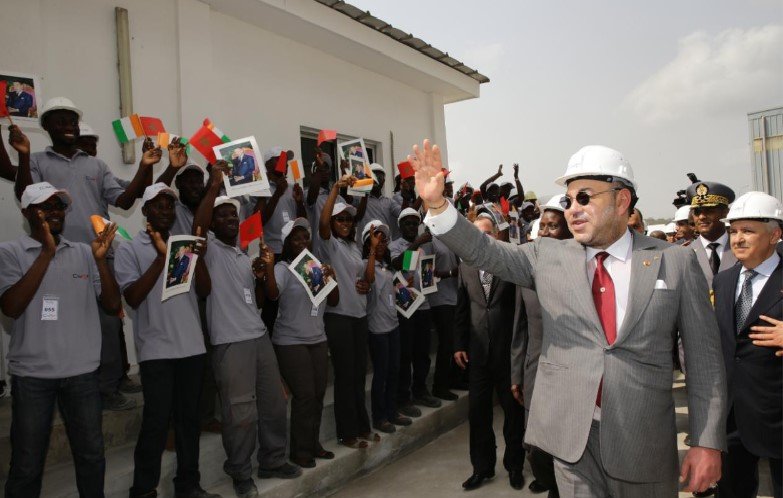 L'image montre un groupe de personnes, probablement des travailleurs ou des étudiants, levant des affiches avec des photos tout en saluant une personne qui semble être un dirigeant. Ce dernier porte un casque de sécurité et coûte en costume, indiquant une visite d'inspection ou une inauguration. Les personnes en arrière-plan sont toutes habillées de manière uniforme et semblent enthousiastes, créant une atmosphère de célébration. L'environnement est extérieur, avec un bâtiment en arrière-plan.