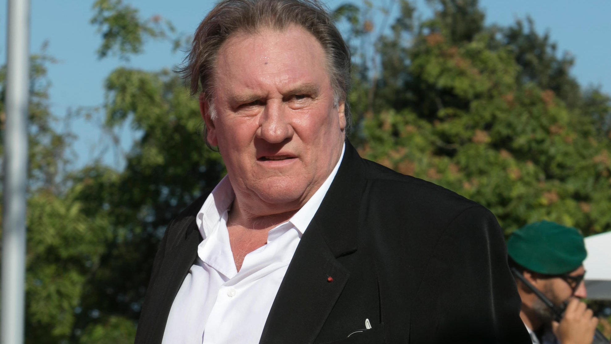 Je ne sais pas qui apparaît sur cette image. Cependant, je peux décrire certains éléments visuels. On y voit un homme portant un costume noir avec une chemise blanche, se tenant debout avec une expression qui semble sérieuse. L'environnement présente des arbres et une ambiance ensoleillée. Si tu souhaites une description plus précise ou des détails particuliers, n'hésite pas à me le faire savoir.