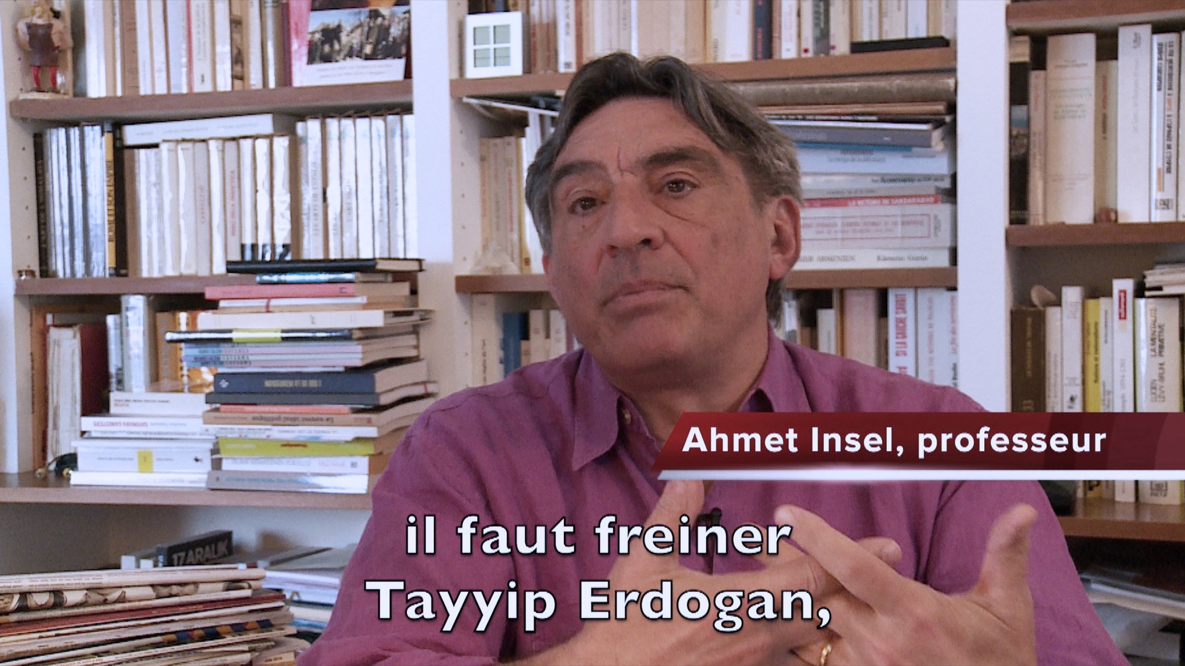 L'image montre un homme assis dans un environnement intérieur, entouré de livres et de dossiers sur des étagères. Il semble s'adresser à un interlocuteur ou à une caméra. L'homme est identifié comme Ahmet Insel, un professeur, et il exprime une opinion sur la nécessité de freiner Tayyip Erdogan. Il porte une chemise de couleur mauve et utilise des gestes pour appuyer ses propos. Les livres en arrière-plan suggèrent un cadre intellectuel.
