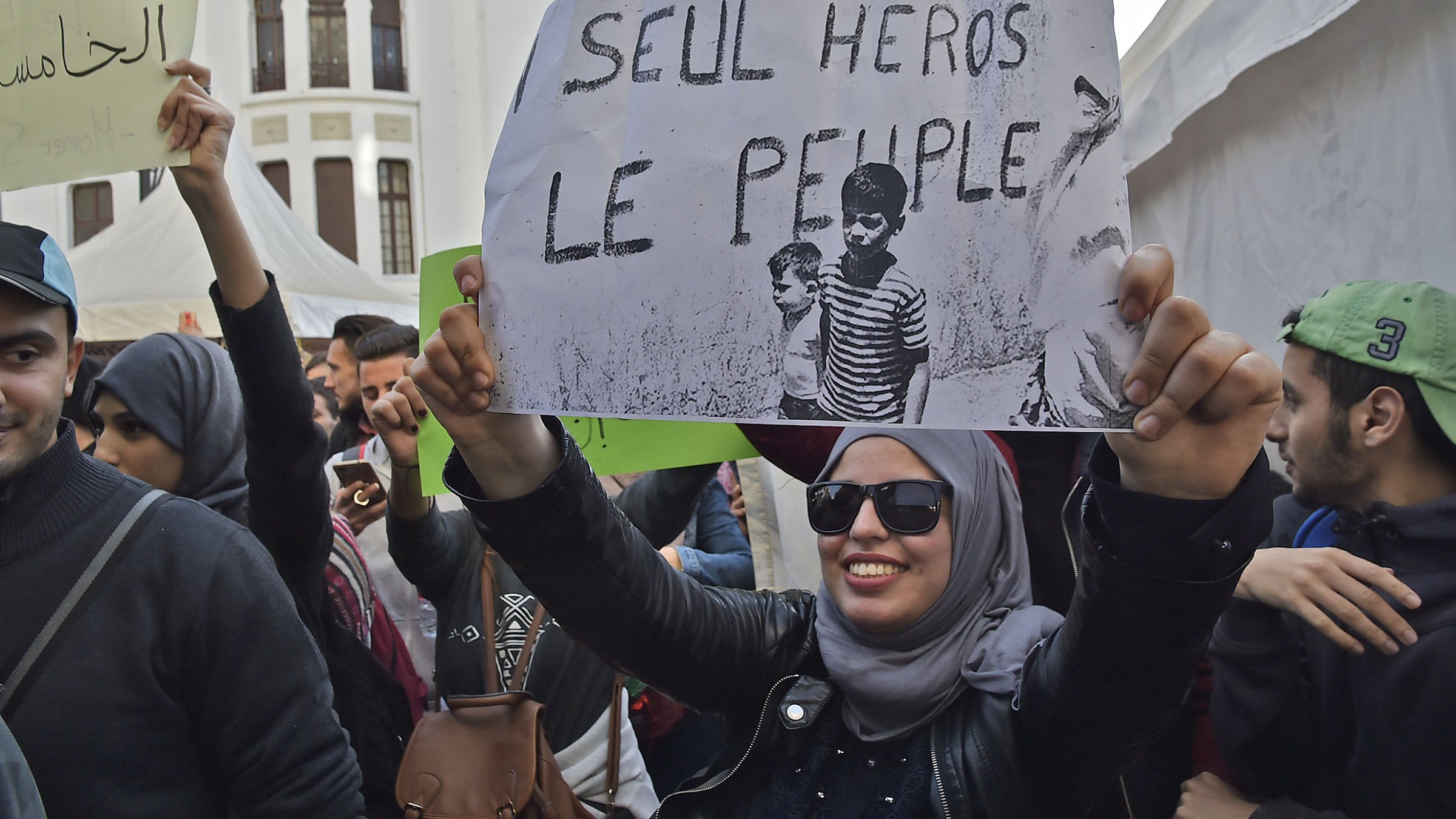 L'image montre une manifestation avec des personnes rassemblées. Au premier plan, une femme portant un hijab et des lunettes de soleil tient une pancarte qui affiche le slogan « SEUL HÉROS LE PEUPLE ? ». L'ambiance semble être engagée, avec d'autres manifestants en arrière-plan et des banderoles visibles. Le décor pourrait suggérer une mobilisation sociale ou politique.
