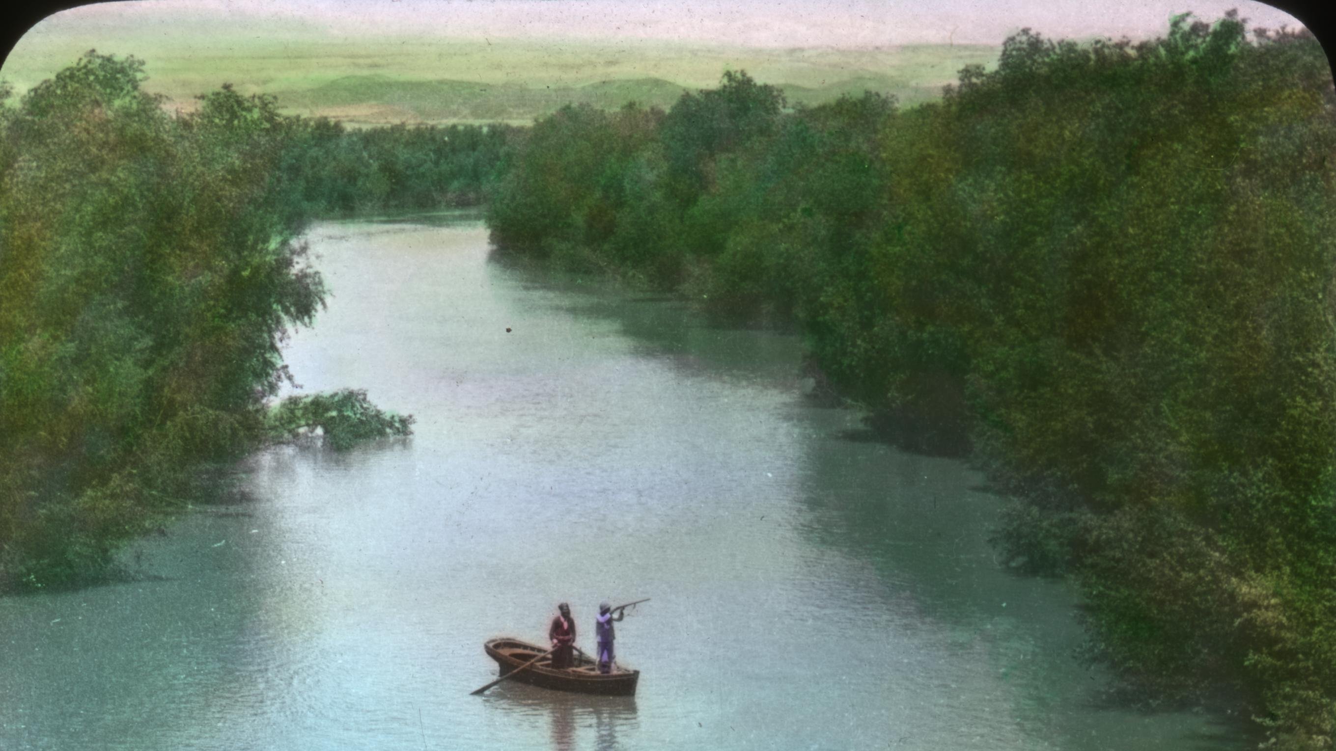 L'image montre une scène paisible au bord d'une rivière. Deux personnes se tiennent sur la berge, regardant une barque qui navigue sur l'eau. La barque semble être occupée par deux personnes qui sont en train de pêcher ou d'observer les environs. La végétation entourant la rivière est dense et verdoyante, ajoutant une touche de nature à la scène. Le ciel est clair et dégage une atmosphère calme et sereine.