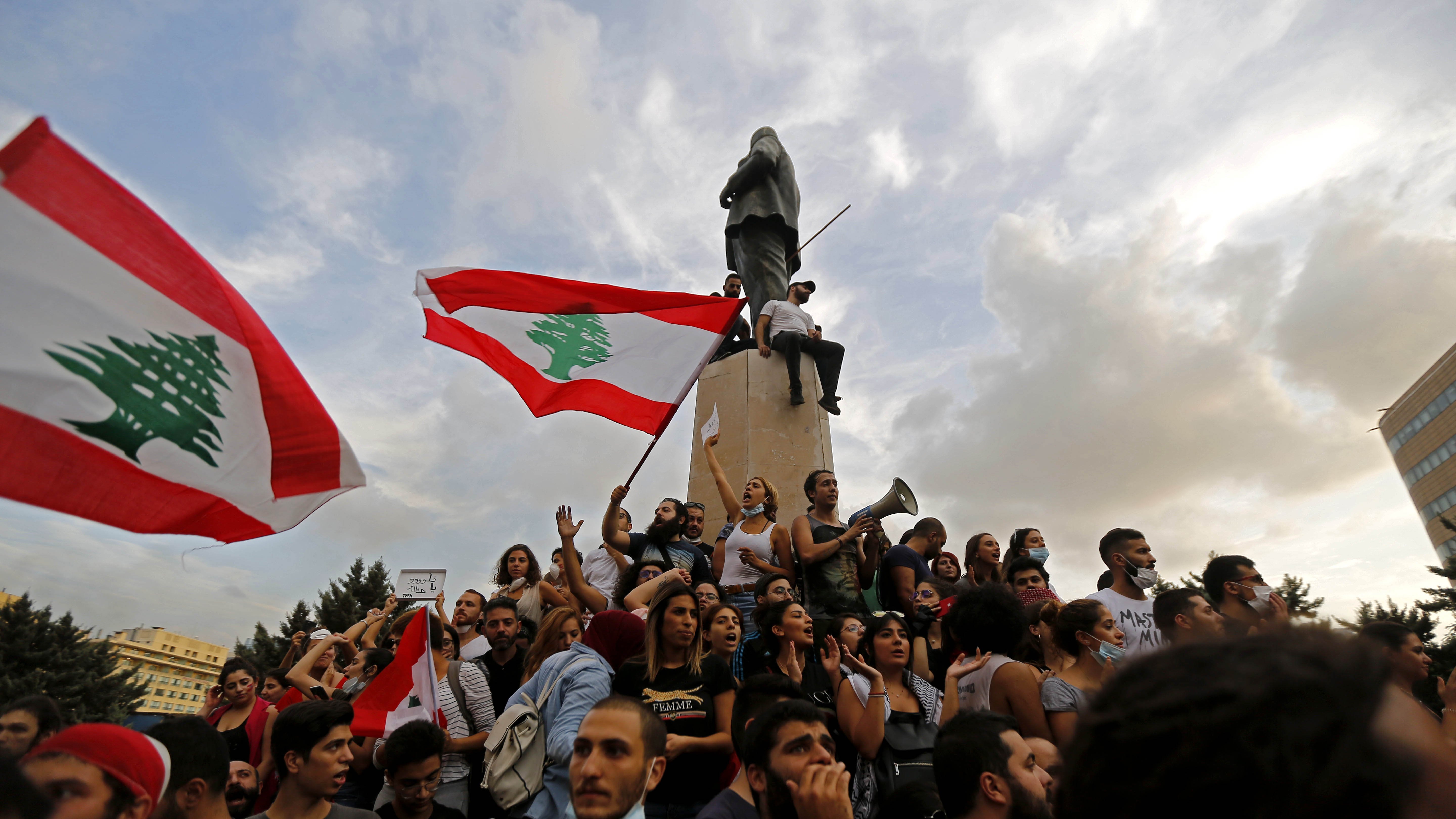 L'image montre une manifestation au Liban. Des groupes de personnes sont rassemblés autour d'un monument, tenant des drapeaux libanais. Certains manifestants chantent ou s'expriment à l'aide de mégaphones. L'atmosphère semble dynamique et engagée, avec des nuages dans le ciel qui ajoutent une ambiance dramatique à la scène.