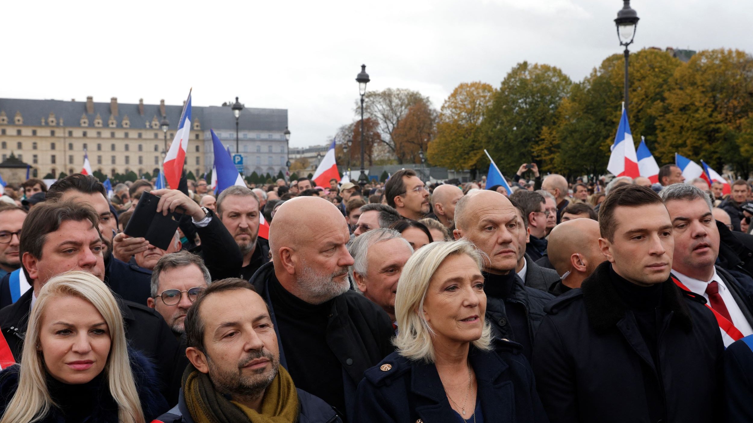 L'image montre une foule rassemblée lors d'un événement en plein air. Au premier plan, plusieurs personnes sont visibles, dont certaines portent des écharpes tricolores aux couleurs du drapeau français. On aperçoit également des drapeaux français agités par des membres de la foule. L'arrière-plan présente des bâtiments historiques et un ciel nuageux, ce qui donne une atmosphère de mobilisation. Les expressions des personnes semblent sérieuses et engagées, témoignant de l'importance de l'événement.