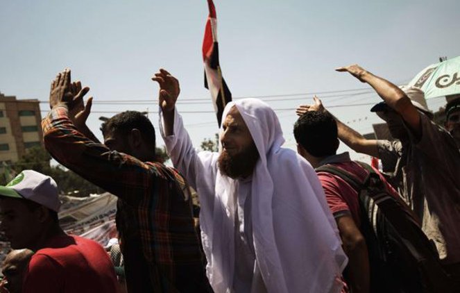 L'image montre une scène de rassemblement ou de manifestation. On peut voir un groupe de personnes levant les bras, probablement en signe de protestation ou de célébration. Parmi eux, un homme se distingue par sa tenue traditionnelle, portant un habit clair et un foulard, tout en brandissant un drapeau. L'ambiance semble être celle d'une grande mobilisation, avec des expressions de détermination ou d'engagement sur les visages des participants. Des bâtiments sont visibles en arrière-plan, suggérant que l'événement se déroule en milieu urbain.