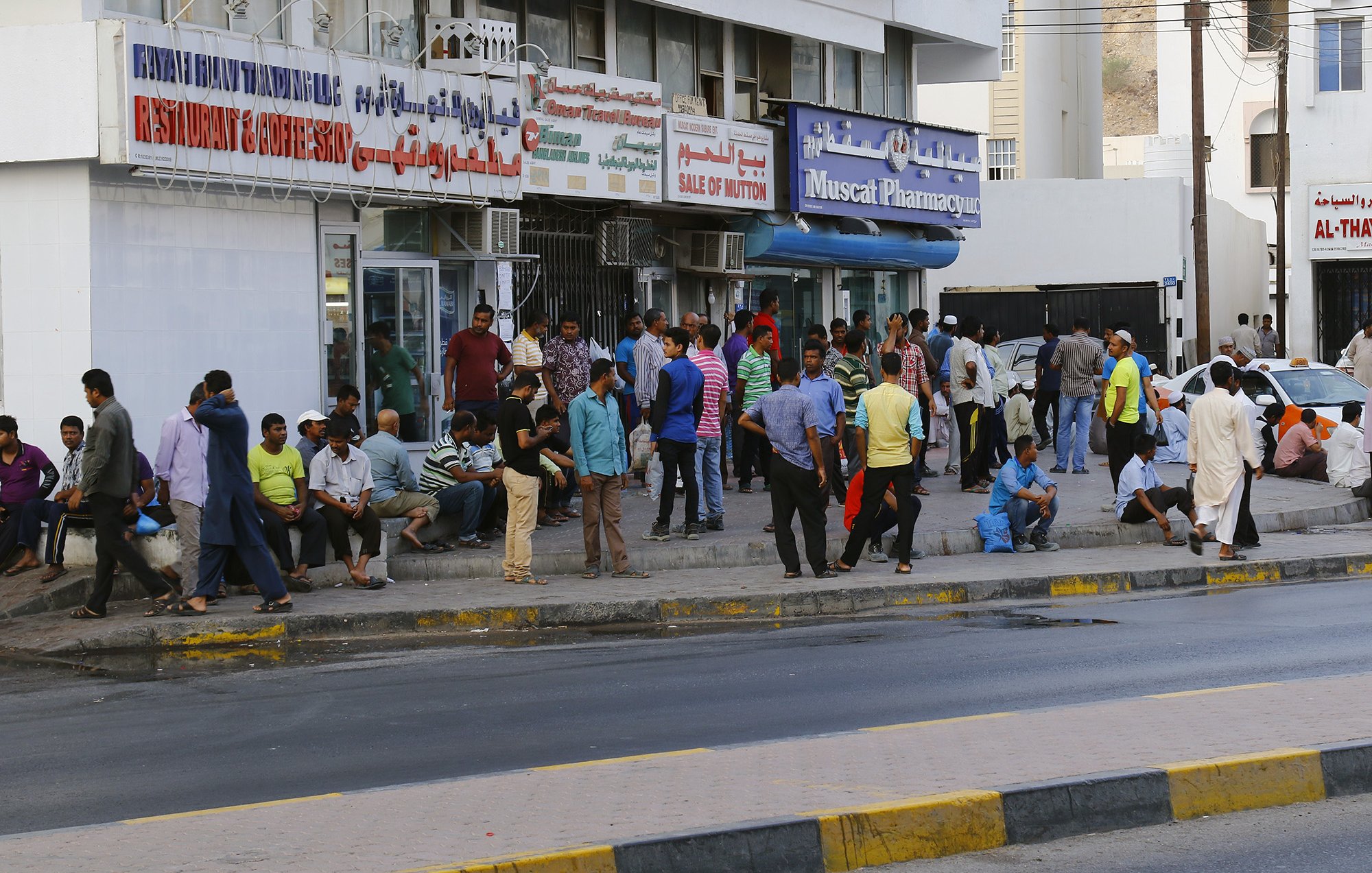 L'image montre une scène urbaine animée, avec un groupe de personnes rassemblées devant un bâtiment. On peut voir des hommes de différentes tenues discutant, assis ou se tenant debout sur le trottoir. Le bâtiment porte des enseignes de restaurants et de pharmacie, indiquant une ambiance commerçante. La route est visible au premier plan, avec une bande jaune délimitant la chaussée. L'atmosphère semble vivante, typique des zones urbaines fréquentées.