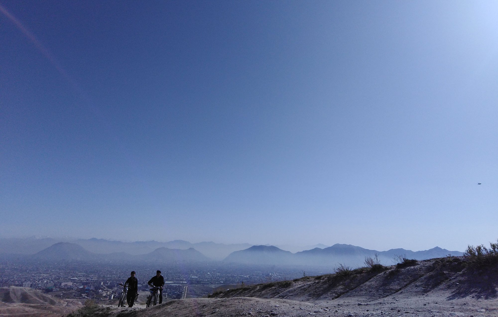 L'image montre deux personnes se tenant sur une colline, contemplant un vaste paysage sous un ciel bleu clair. On peut voir des montagnes à l'horizon et une ville en contrebas, légèrement obscurcie par une brume. La lumière est douce, créant une ambiance paisible et tranquille. Les silhouettes des personnes ajoutent une note humaine à cette scène naturelle.