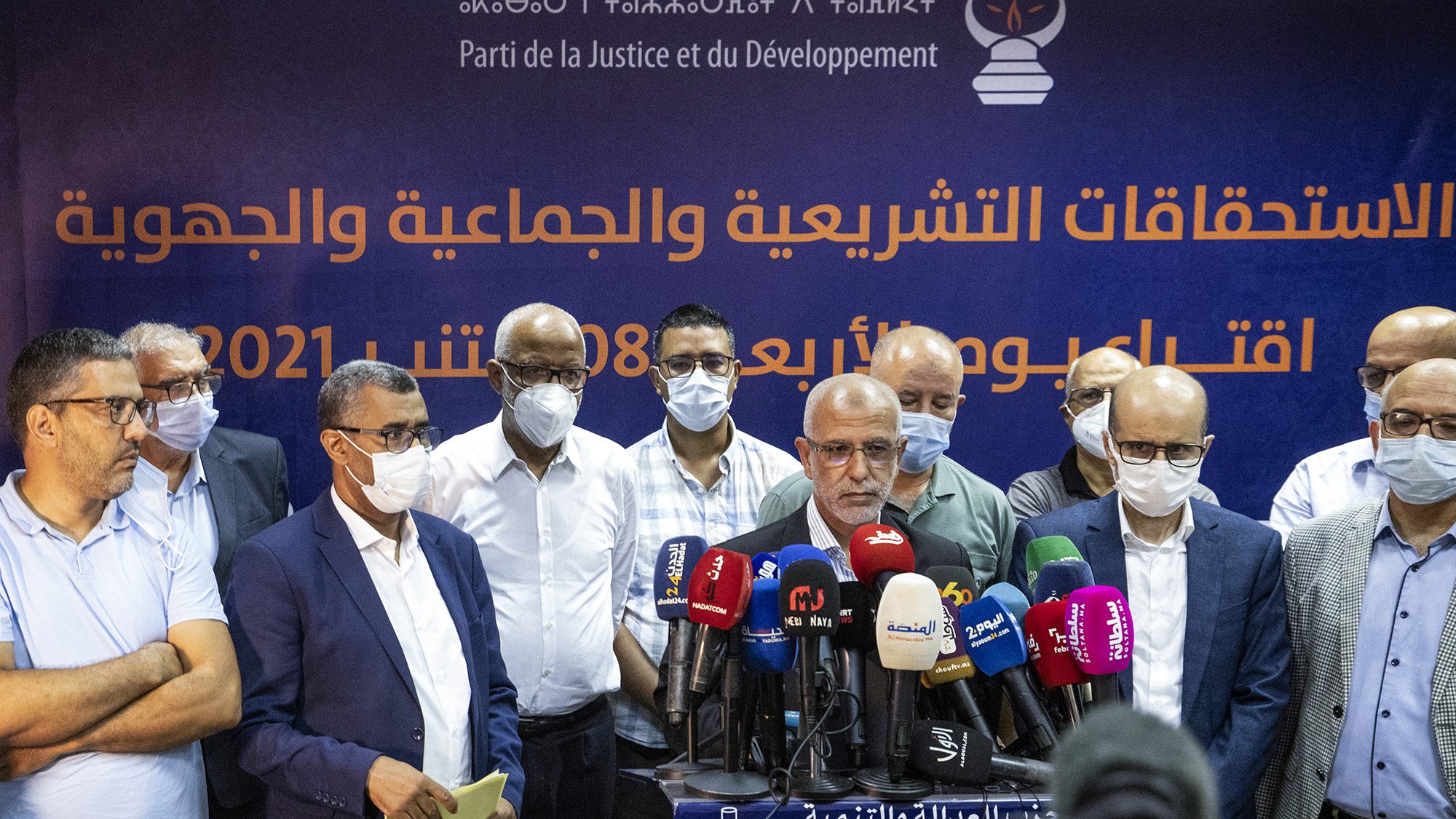 Sur l'image, un groupe d'hommes se tient devant une bannière affichant le nom d'un parti politique, le Parti de la Justice et du Développement. Les hommes portent des masques de protection, ce qui suggère un contexte de santé publique. Ils semblent s'apprêter à faire une déclaration ou une conférence de presse, entourés de microphones et de caméras. L'atmosphère semble sérieuse et professionnelle.