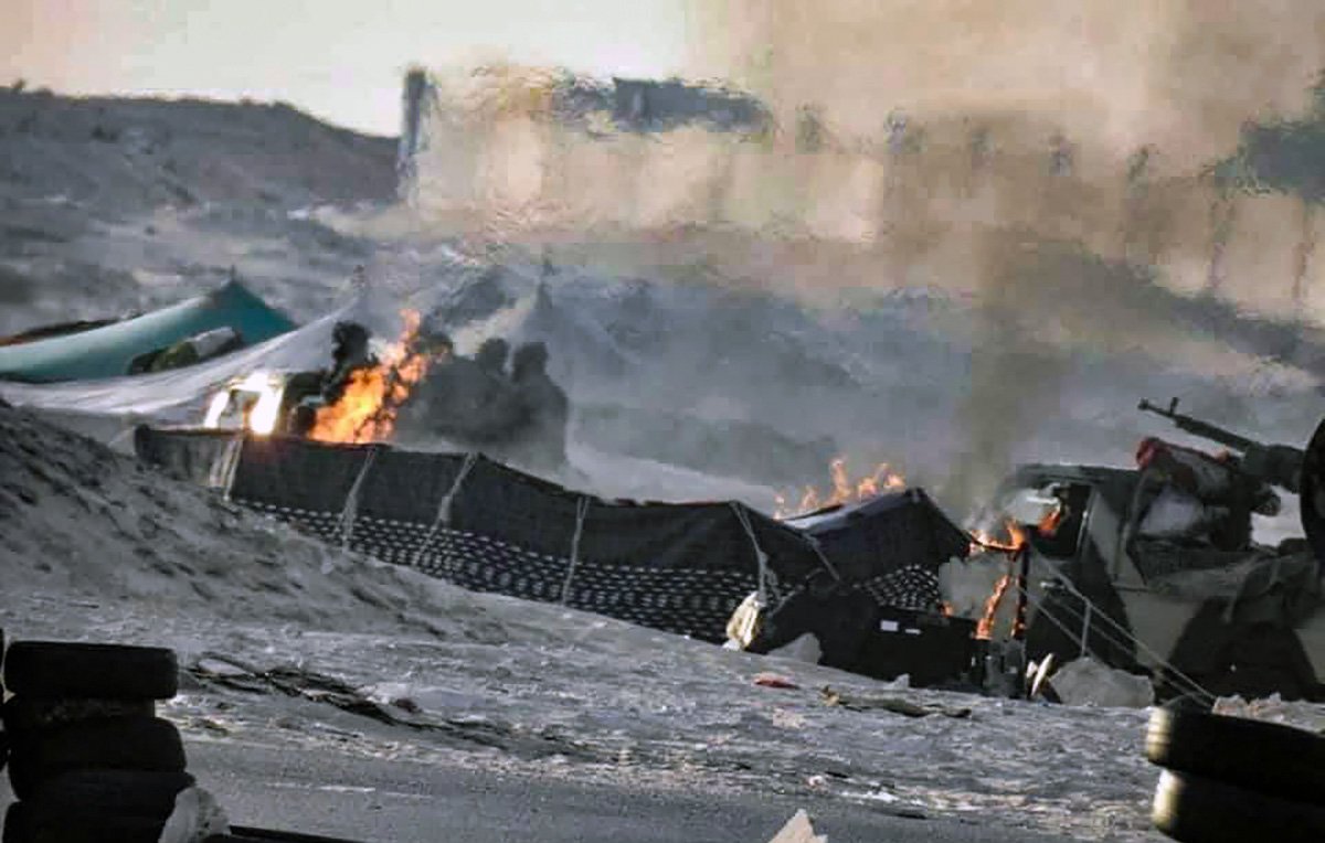 L'image semble représenter une scène de conflit ou de guerre, avec des flammes visibles et de la fumée s'élevant dans l'air. On peut distinguer des structures endommagées et une ambiance chaotique. Le sol est recouvert de débris, et des véhicules militaires ou des équipements peuvent être présents. L'environnement semble aride, suggérant un paysage désertique.