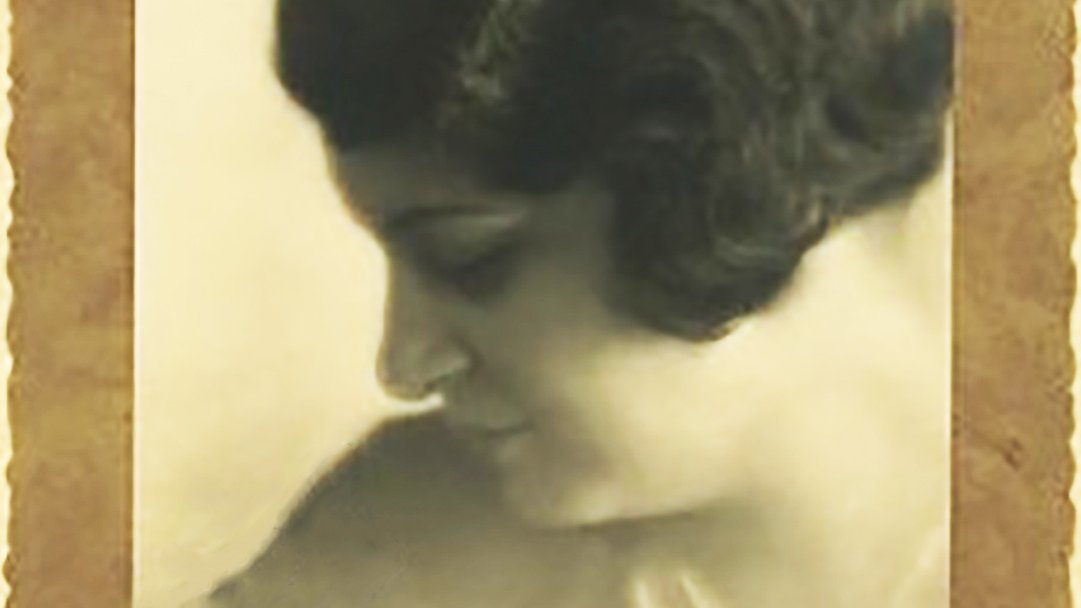 L'image représente un portrait en noir et blanc d'une femme, pris de profil. Elle a les cheveux bouclés et une coiffure élégante typique des années 1920-1930. Son expression est douce et pensive, et elle semble regarder vers le bas, créant une ambiance contemplative. Le fond de l'image est flou, mettant en valeur son visage, et le tout est encadré par une bordure décorative qui ajoute une touche vintage à la photo.