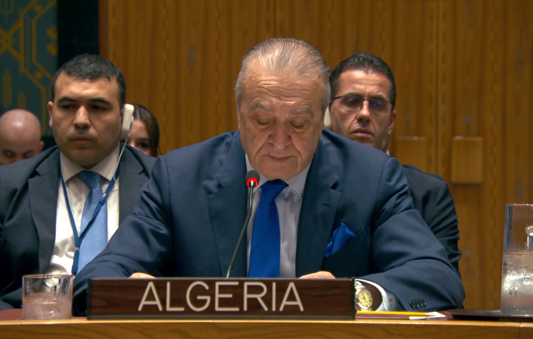 Un homme en costume parle à un podium, des personnes écoutent derrière lui.