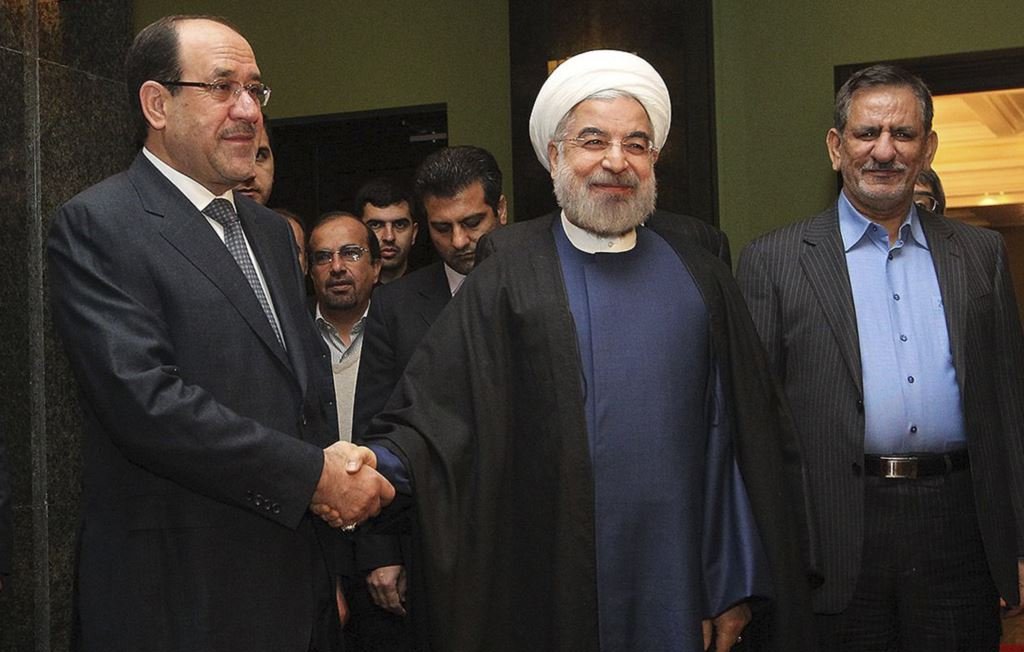 L'image montre trois hommes se tenant debout, avec un homme au centre qui sourit et porte une robe traditionnelle, tandis que les deux autres sont en costume. Ils semblent être en train de se saluer, probablement lors d'une rencontre officielle. L'environnement semble formel, probablement dans un hôtel ou un bâtiment gouvernemental, avec des personnes en arrière-plan.