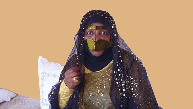 L'image montre une femme vêtue d'une tenue traditionnelle ornée de motifs et de broderies. Elle porte un voile noir qui couvre son visage en grande partie, laissant seulement ses yeux visibles, ce qui ajoute une touche de mystère. Sa robe est richement décorée, avec des couleurs sombres et des embellissements scintillants. À côté d'elle, on aperçoit des objets traditionnels, ce qui suggère une ambiance culturelle. L'arrière-plan est d'une couleur unie, mettant en valeur sa présence.