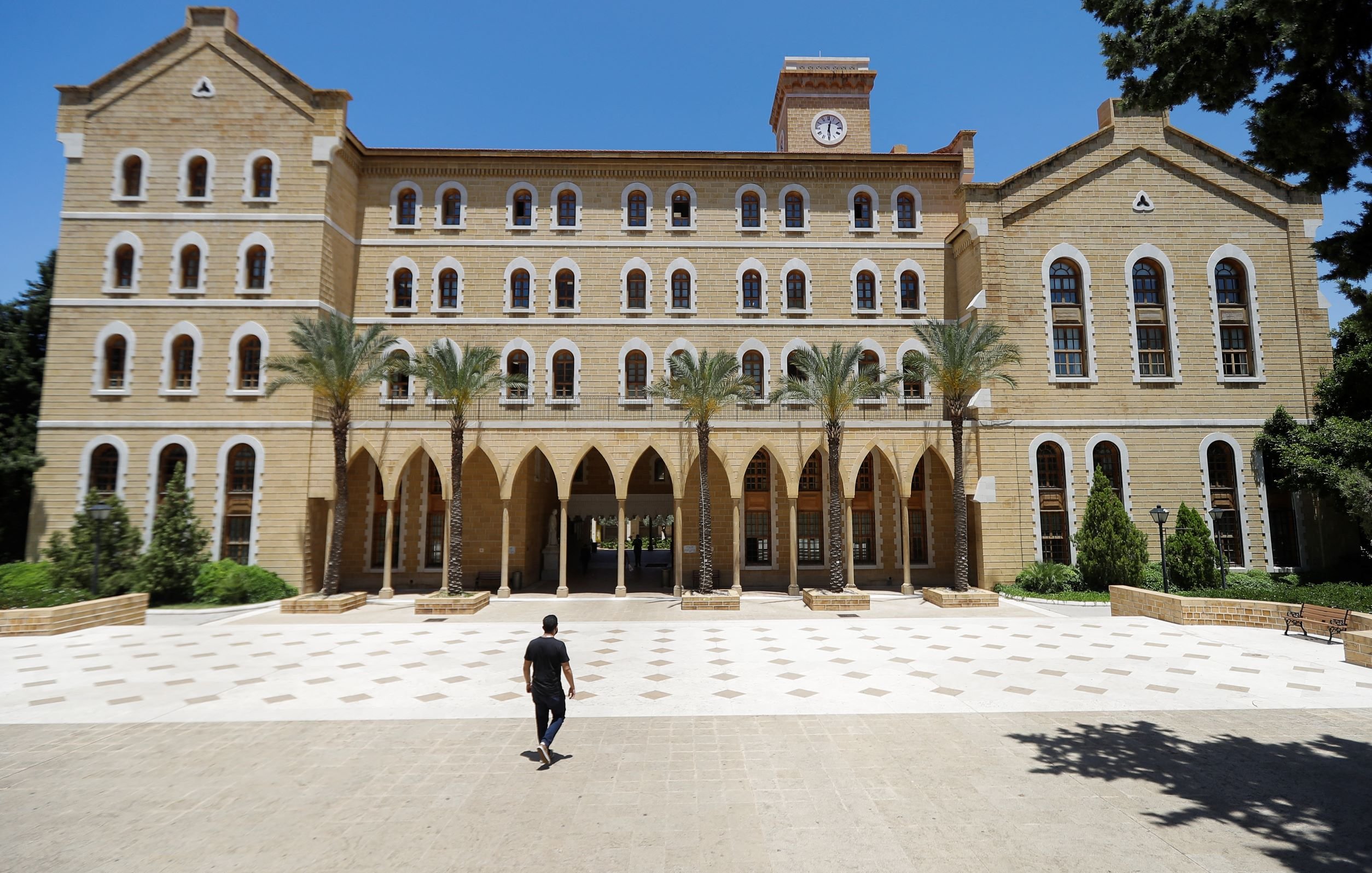 L'image montre un grand bâtiment en pierre avec une architecture traditionnelle, comportant plusieurs fenêtres à arc et des colonnes. Au premier plan, une personne marche seule sur une place pavée, tandis que des palmiers sont visibles sur les côtés. Le ciel est clair et bleu, ce qui donne une atmosphère ensoleillée. Le bâtiment semble être une institution éducative ou un lieu historique.
