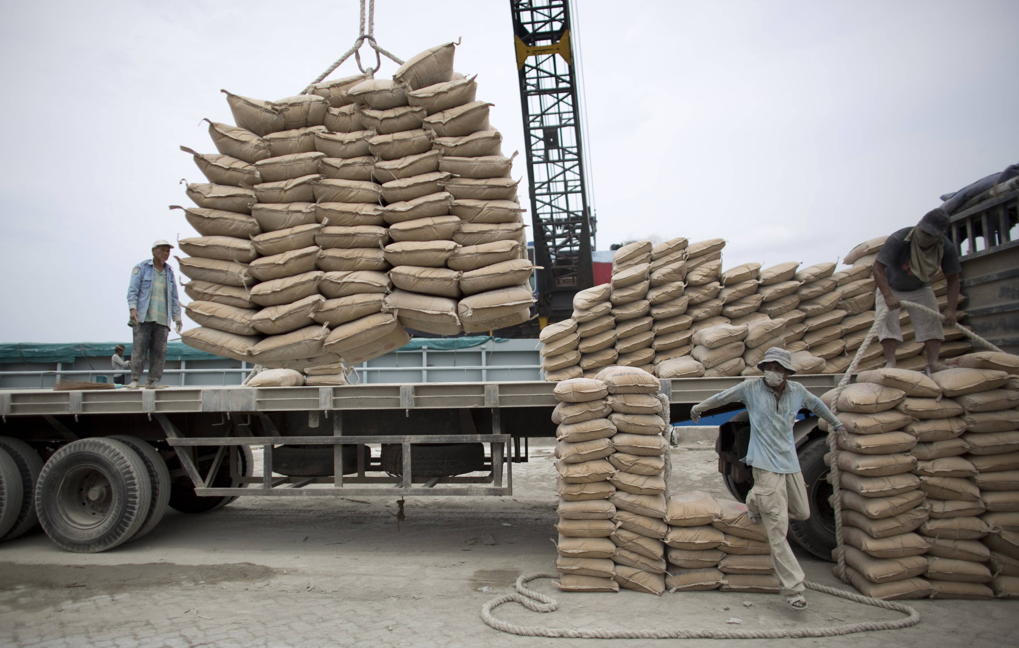 L'image montre une scène de travail au port ou dans un chantier. On y voit des hommes qui manipulent des sacs de sable ou de ciment. Certains sont en train de charger des sacs sur un camion à l'aide d'une grue, tandis que d'autres semblent organiser les sacs déjà empilés. L'environnement est industriel, avec des matériaux de construction et des équipements lourds visibles en arrière-plan. Le ciel est nuageux, ce qui donne une ambiance un peu grise à la scène.