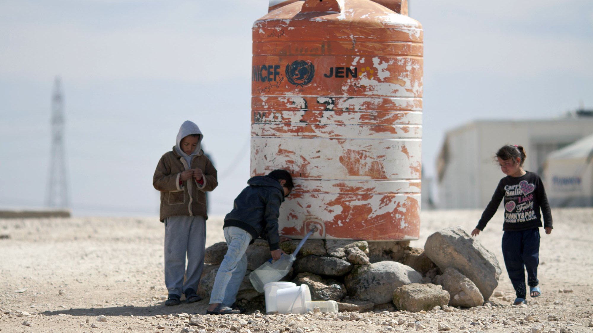 L'image montre un groupe d'enfants autour d'un réservoir d'eau. Le réservoir, de couleur orange et marqué par des signes d'usure, semble abriter l'eau potable. Les enfants, dont l'un est en train de remplir un récipient, sont habillés de manière simple et semblent se trouver dans un environnement désertique. On peut voir des pierres autour du réservoir, et le paysage en arrière-plan est sobre, suggérant une situation de besoin en ressources.