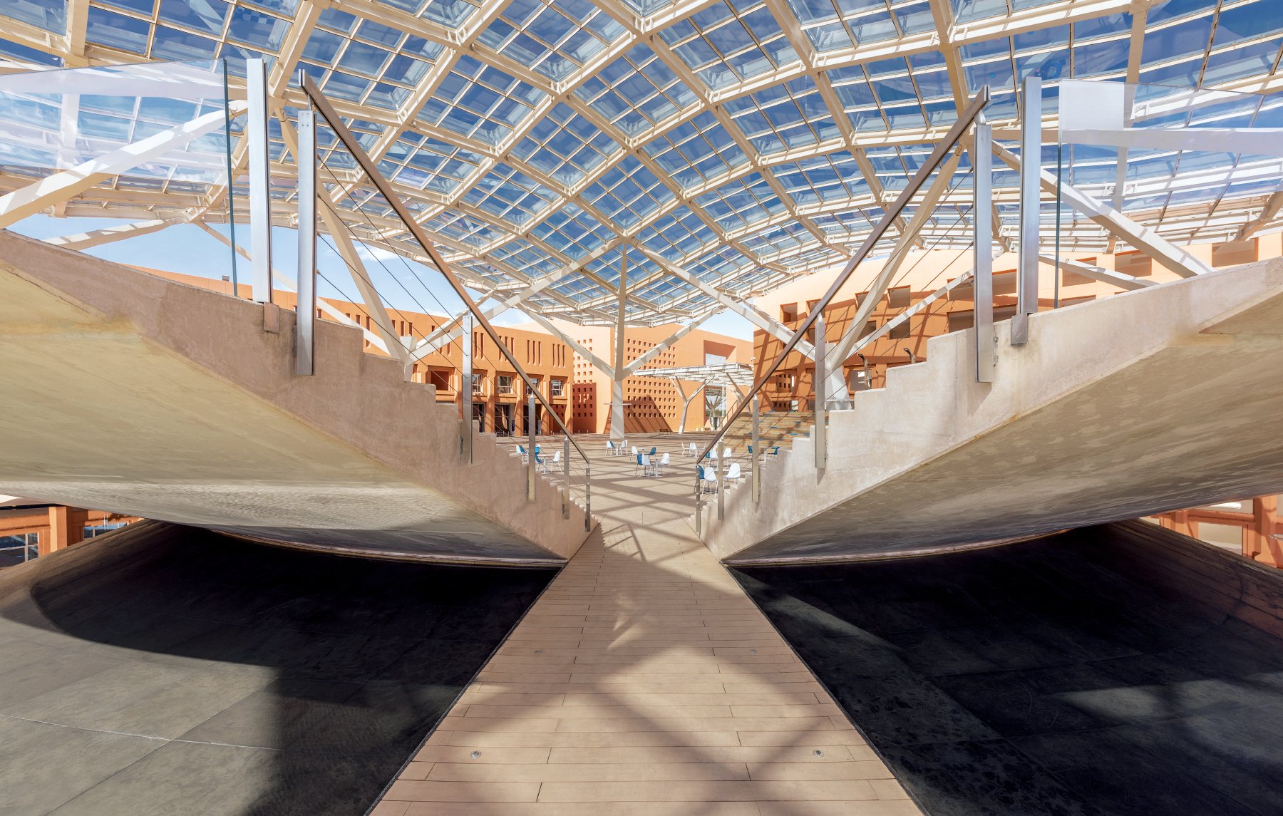L'image montre un intérieur architectural moderne avec une structure impressionnante. Le plafond est en verre, permettant à la lumière naturelle d'entrer, et crée une ambiance lumineuse. Les murs et les surfaces sont en béton et en bois, offrant un contraste intéressant. Des passerelles en bois traversent l'espace, menant à des zones ouvertes. La conception suggère un espace fonctionnel et esthétique, propice à des activités variées.
