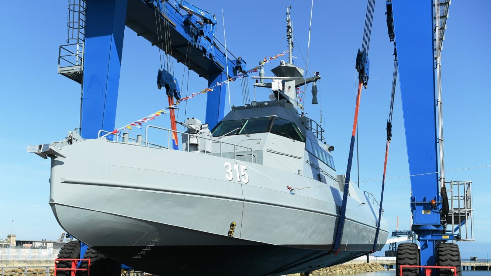 L'image montre un navire militaire en cours de manutention. Il est positionné sur un quai, soutenu par des grues bleues qui le soulèvent avec des sangles. Le bateau est de couleur grise et porte le numéro 315. Le ciel est clair, suggérant une journée ensoleillée. Cette scène évoque des opérations de maintenance ou de construction navale.