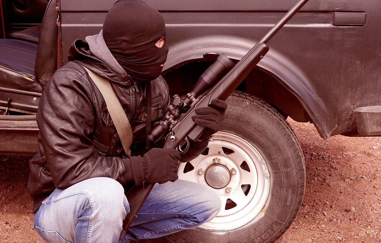 L'image représente une personne en position accroupie, portant un masque noir qui couvre son visage. Elle est habillée d'une veste en cuir et de gants noirs. Dans ses mains, elle tient un fusil équipé d'un accessoire de visée. En arrière-plan, on peut voir une camionnette avec une roue visible, suggérant un environnement extérieur. L'ensemble dégage une atmosphère tendue et dramatique.