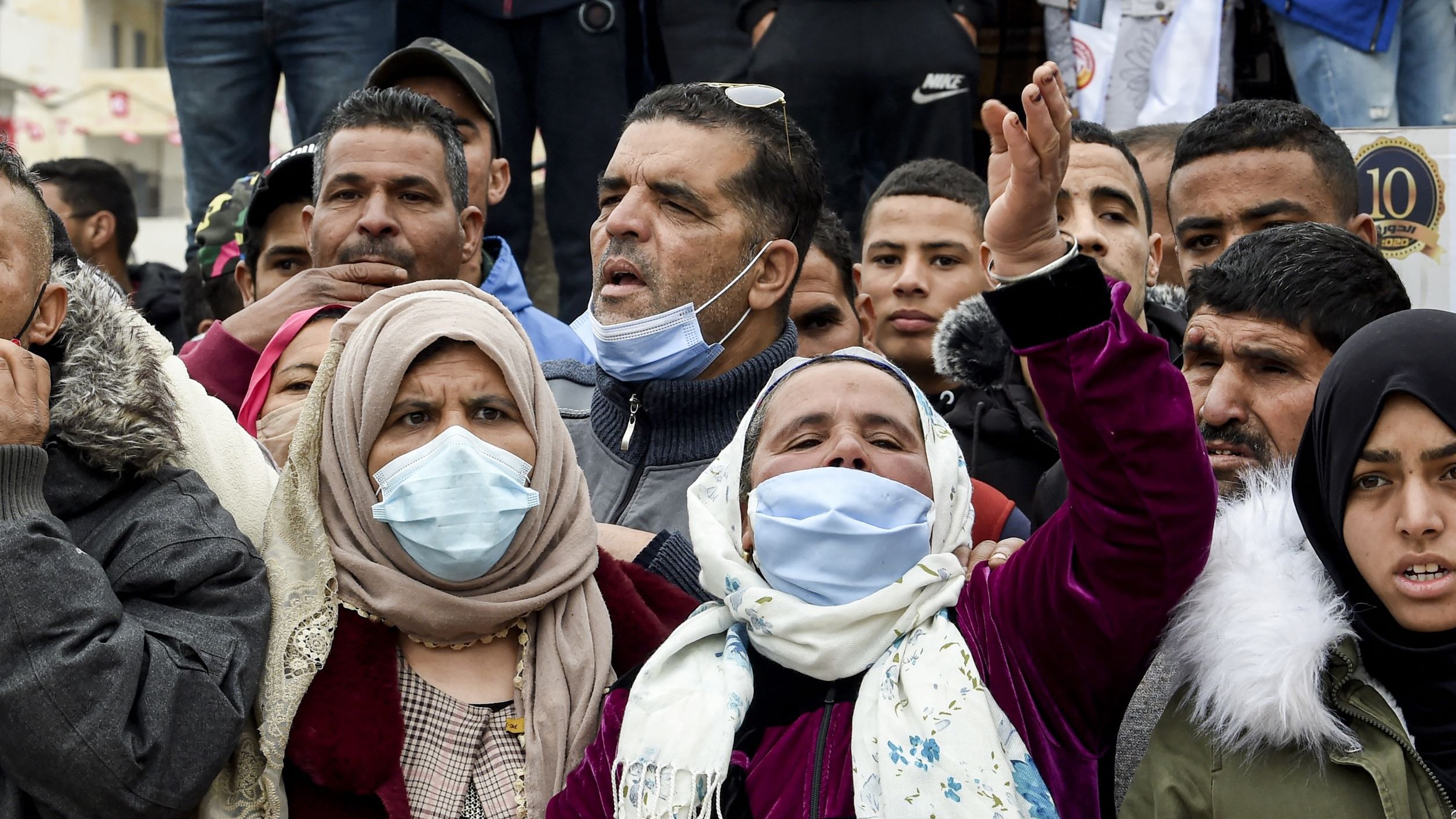 L'image montre une foule de personnes, principalement des hommes et des femmes, réunies pour exprimer leurs émotions. Certaines personnes portent des masques faciaux, et il y a des expressions de détermination et de protestation parmi les membres du groupe. Les vêtements sont variés, avec des accents de couleurs différentes. L'ambiance semble animée et engagée, suggérant une manifestation ou un rassemblement pour une cause.