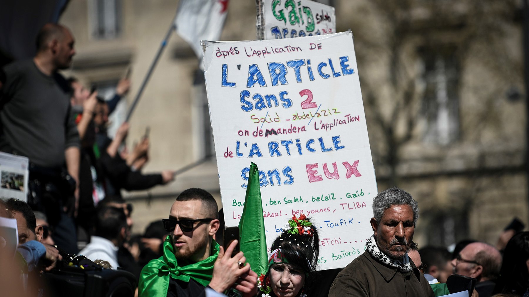 L'image montre une manifestation avec des participants tenant des pancartes. L'une des pancartes est bien visible et contient du texte en français, notamment une référence à l'article 2 et des demandes concernant cet article. Les manifestants portent des vêtements aux couleurs de l'Algérie, ce qui suggère un lien avec des préoccupations politiques ou sociales. L'atmosphère semble sereine mais déterminée, avec des gens rassemblés pour exprimer leurs revendications.