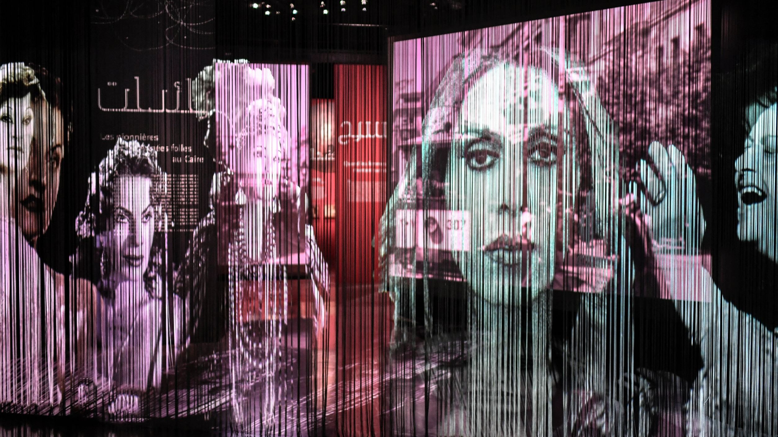 L'image présente une installation artistique composée de plusieurs grands panneaux transparents sur lesquels sont projetées des images en noir et blanc de visages féminins. Les projections créent un effet de profondeur et de mouvement, avec des formes et des silhouettes qui se superposent. Le fond est teinté de couleurs douces, suggérant une ambiance à la fois artistique et contemplative. Ce type d'installation évoque une réflexion sur la représentation et l'identité, tout en intégrant des éléments visuels captivants.