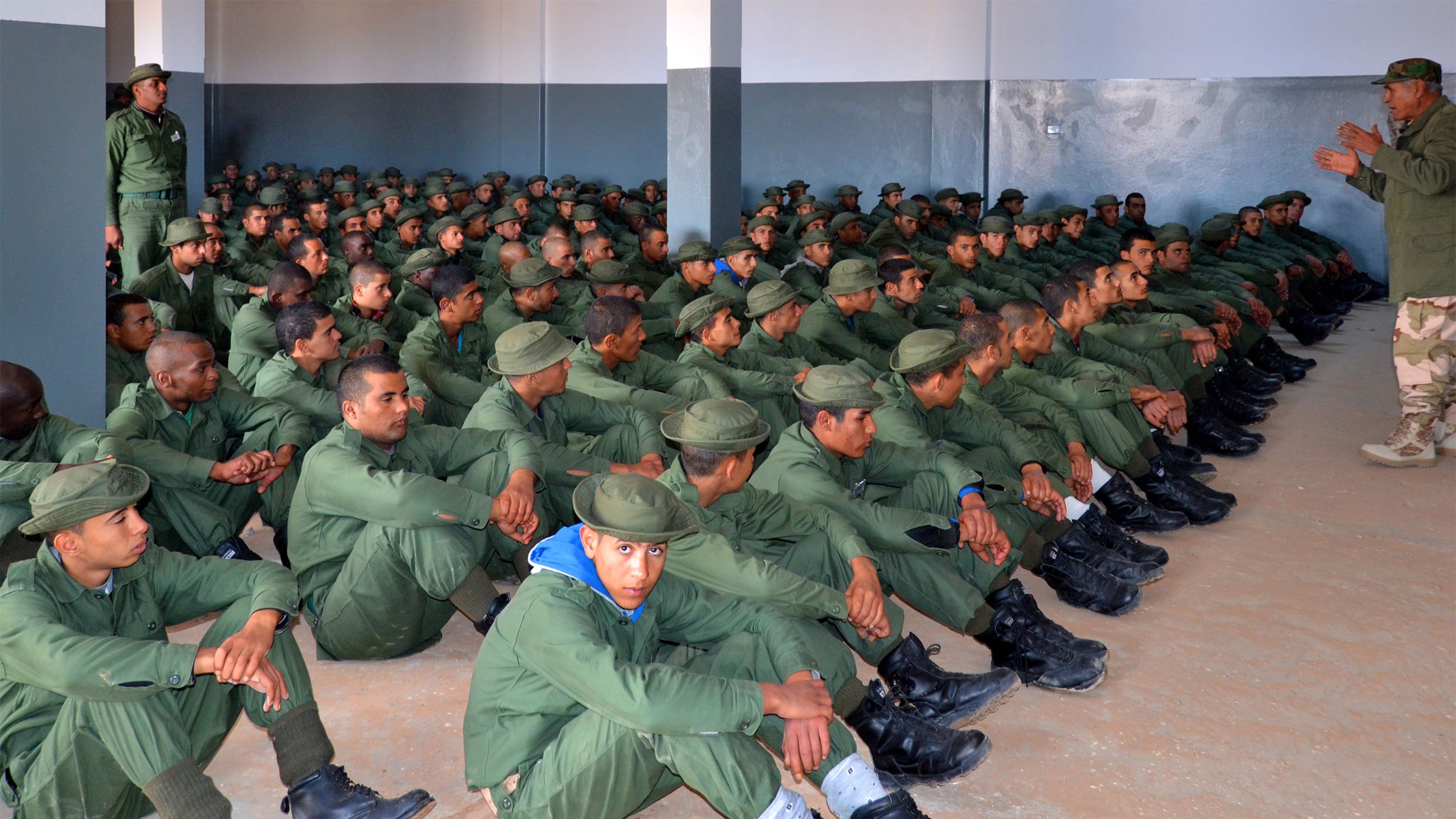 L'image montre un grand groupe de soldats assis dans une salle. Ils portent des uniformes verts et des bottes noires, et sont alignés en rangées sur le sol. Un instructeur, également en uniforme, se tient debout devant eux, semblant donner des instructions ou une formation. Les murs de la salle sont peints en gris, et l'atmosphère semble sérieuse et concentrée.