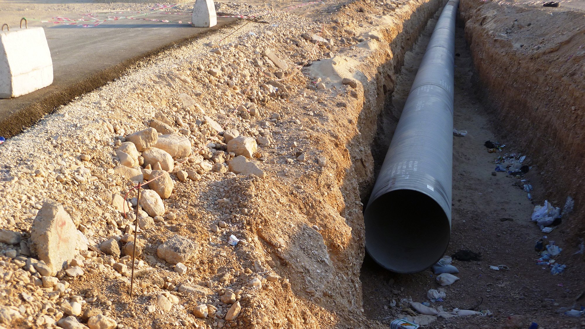 L'image montre un espace de construction où un grand tuyau en métal est disposé dans une tranchée creusée dans le sol. Le terrain environnant est aride et rocailleux, avec quelques débris, comme des bouteilles, dispersés dans la tranchée. Au fond, on peut voir des engins de chantier et une route partiellement construite. L'ambiance générale évoque un projet d'infrastructure en cours.
