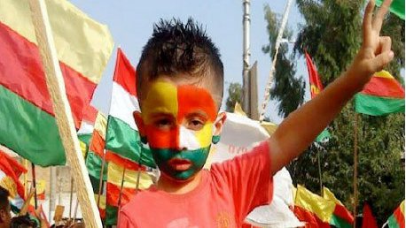 L'image montre un jeune garçon au milieu d'une manifestation ou d'une célébration. Il a le visage peint aux couleurs du drapeau, probablement inspirées par les couleurs du Cameroun, avec des nuances de vert, rouge et jaune. Il porte un t-shirt rouge et affiche un signe de paix avec sa main, tout en étant entouré de drapeaux aux couleurs similaires. L'atmosphère semble festive et engagée.