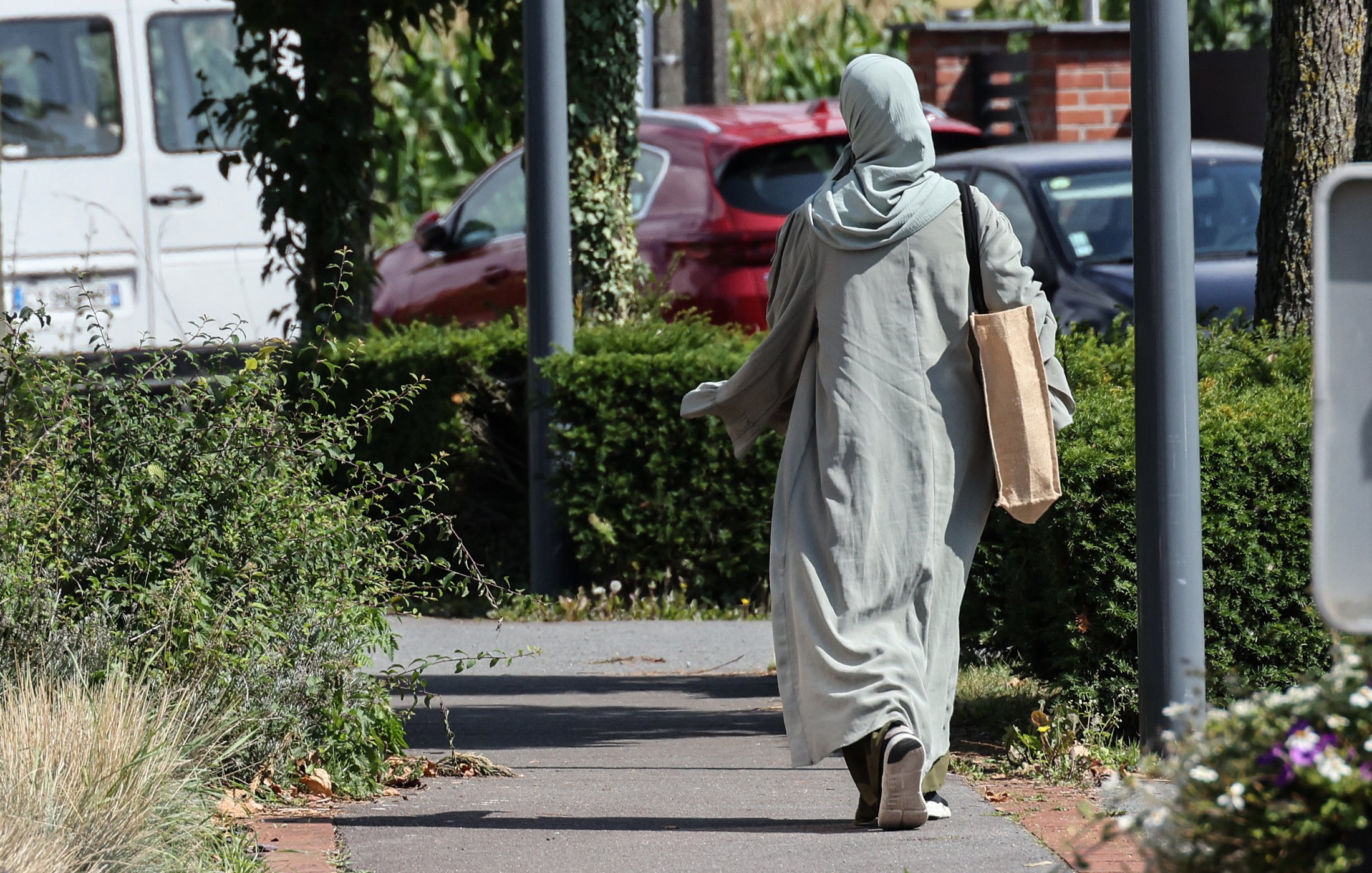 L'image montre une personne marchant sur un chemin. Elle porte une longue robe de couleur claire et un foulard. Elle tient un grand sac en papier dans une main. Le décor environnant inclut des buissons et des arbres, avec des voitures en arrière-plan. La scène semble tranquille et ensoleillée.