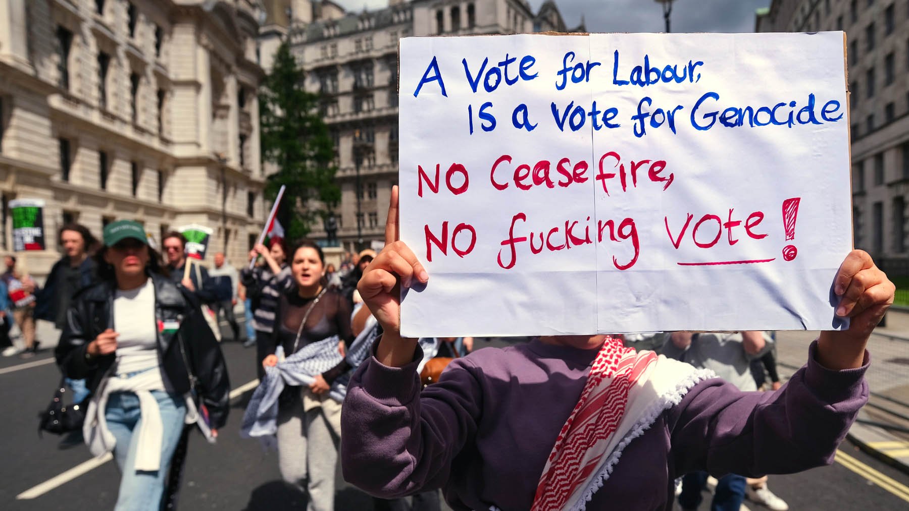 L'image montre une manifestation où des personnes défilent dans la rue, tenant des pancartes. Au premier plan, une personne tient une grande affiche avec un message fort. La pancarte indique que voter pour un certain parti est associé à des accusations graves, et elle appelle à ne pas voter sans un cessez-le-feu. Les manifestants semblent passionnés et engagés, exprimant leur désaccord par des slogans. L'ambiance est empreinte de tension et d'émotion.