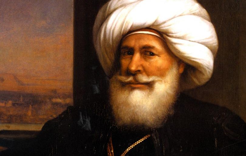 L'image représente un homme portant une tenue traditionnelle, avec un turban distinctif et une barbe blanche. Il a un sourire visible et semble confiant. L'homme est assis, tenant une épée décorative dans sa main, ce qui suggère une certaine dignité ou autorité. En arrière-plan, on peut apercevoir un paysage, probablement représentant une scène architecturale ou naturelle. L'ensemble donne une impression de grandeur et de culture.