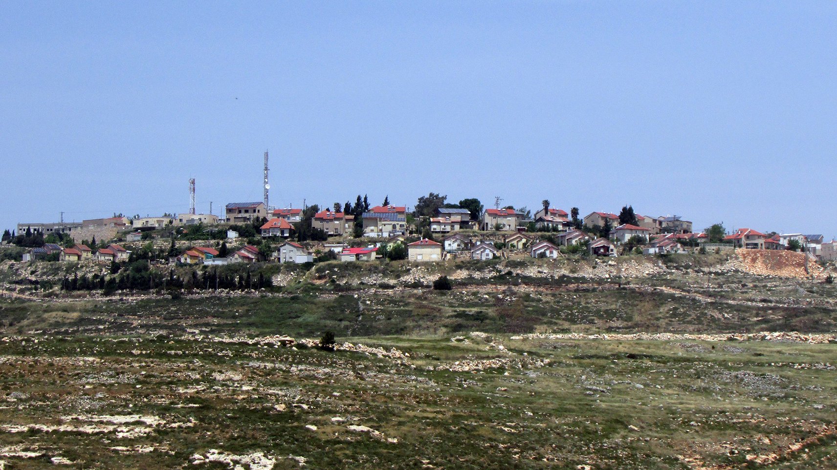 L'image montre un village situé sur une colline, avec des maisons dispersées, certaines avec des toits rouges. On peut également voir des installations comme des antennes de communication. Le paysage environnant est composé de champs et de collines vertes, suggérant une région rurale. Le ciel est clair et bleue, ce qui donne une ambiance tranquille à la scène.
