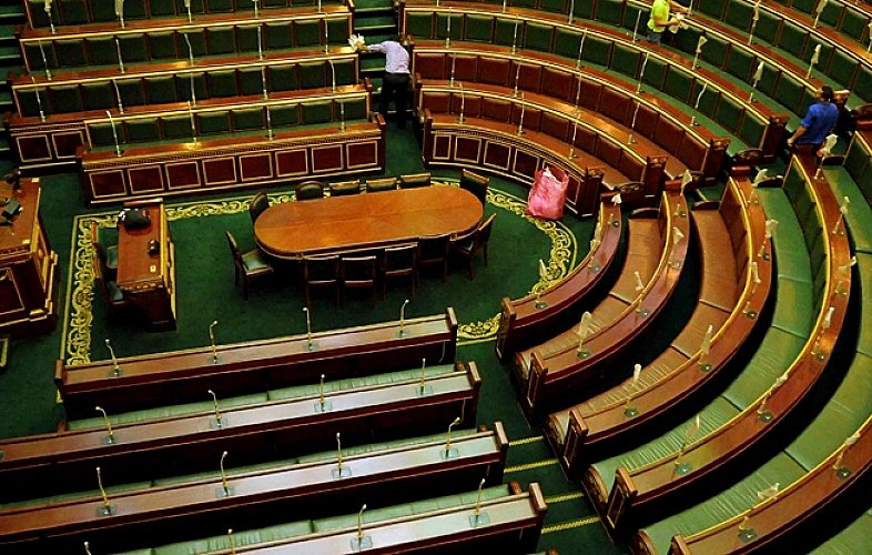 L'image montre l'intérieur d'une assemblée, probablement un parlement ou une salle de réunion officielle. On peut voir des rangées de sièges en bois oblongs disposés en arc de cercle, avec un grand bureau central ovale. Les bancs sont vides, ce qui donne une impression de calme. La décoration est sobre, avec des éléments en bois et un tapis vert. Deux personnes sont visibles, probablement en train de ranger ou d'entretenir l'espace. L'ambiance générale semble formelle et institutionnelle.