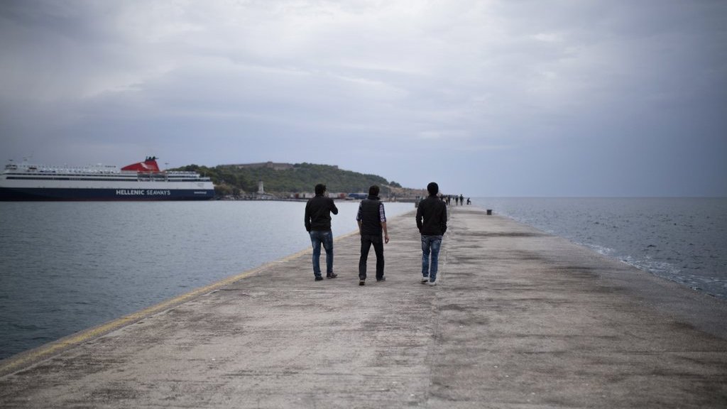 L'image montre trois personnes marchant sur un quai. Le ciel est nuageux, et l'atmosphère est calme. À l'arrière-plan, on peut voir un ferry amarré et une petite île. Le quai est en béton, et l'eau semble calme, reflétant les nuages. Les silhouettes des gens ajoutent une touche de vie à cette scène paisible.