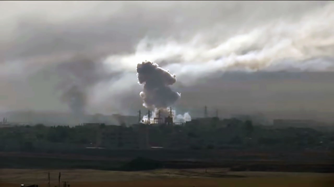 L'image montre une scène dramatique avec des nuages de fumée épais s'élevant dans le ciel, probablement à la suite d'une explosion ou d'un bombardement. On peut voir des silhouettes d'infrastructures industrielles ou urbaines partiellement obscurcies par la fumée. L'atmosphère est sombre et chargée, suggérant des conditions de conflit ou de destruction. La lumière semble faible, ajoutant à l'impression de chaos et de détresse.
