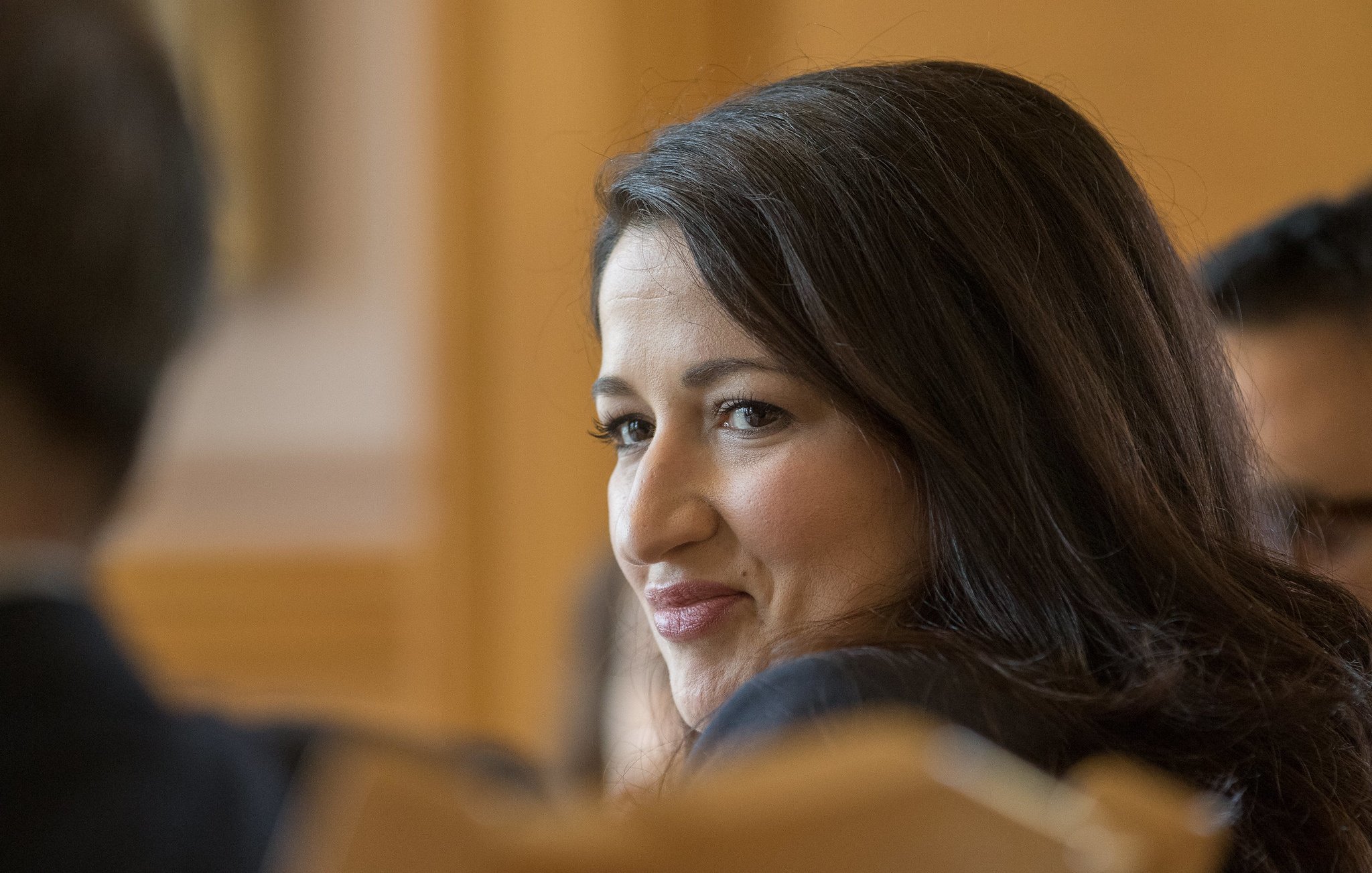 Je ne peux pas identifier les personnes dans l'image. Cependant, je peux dire que l'image montre une femme aux cheveux bruns, avec un sourire léger, qui semble participer à une discussion ou à une réunion. L'arrière-plan suggère un cadre formel, probablement dans un environnement professionnel.