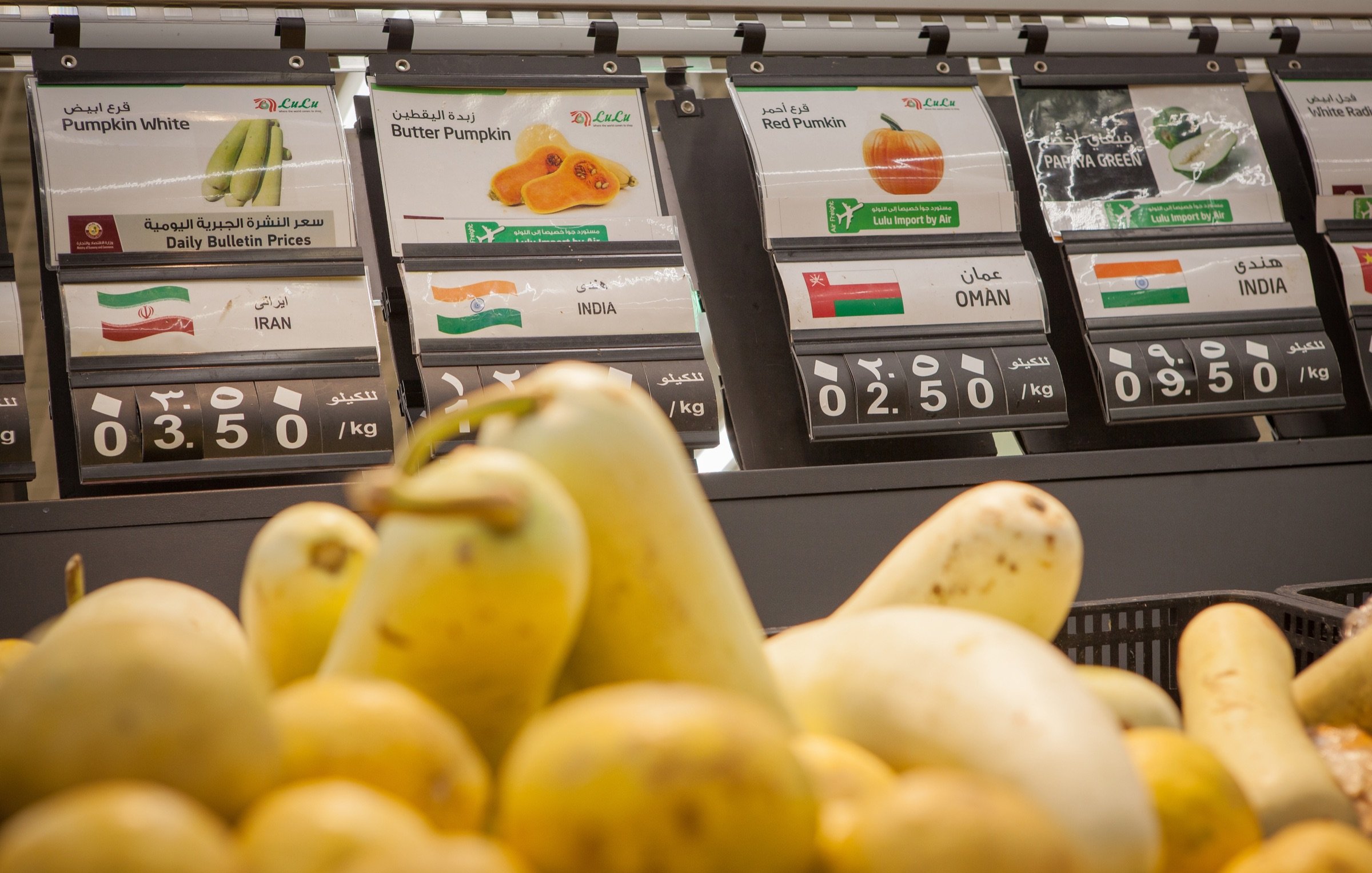 L'image présente un banc de marché dans un magasin, où divers types de courges sont exposés. Au premier plan, on voit des courges de différentes formes et couleurs. En arrière-plan, des étiquettes indiquent les prix et les origines des produits, avec des mentions comme "Courge blanche" et "Courge butternut", ainsi que des pays d'origine tels que l'Iran, Oman et l'Inde. Les prix sont affichés en kilos. L'ensemble crée une ambiance de supermarché frais et coloré.
