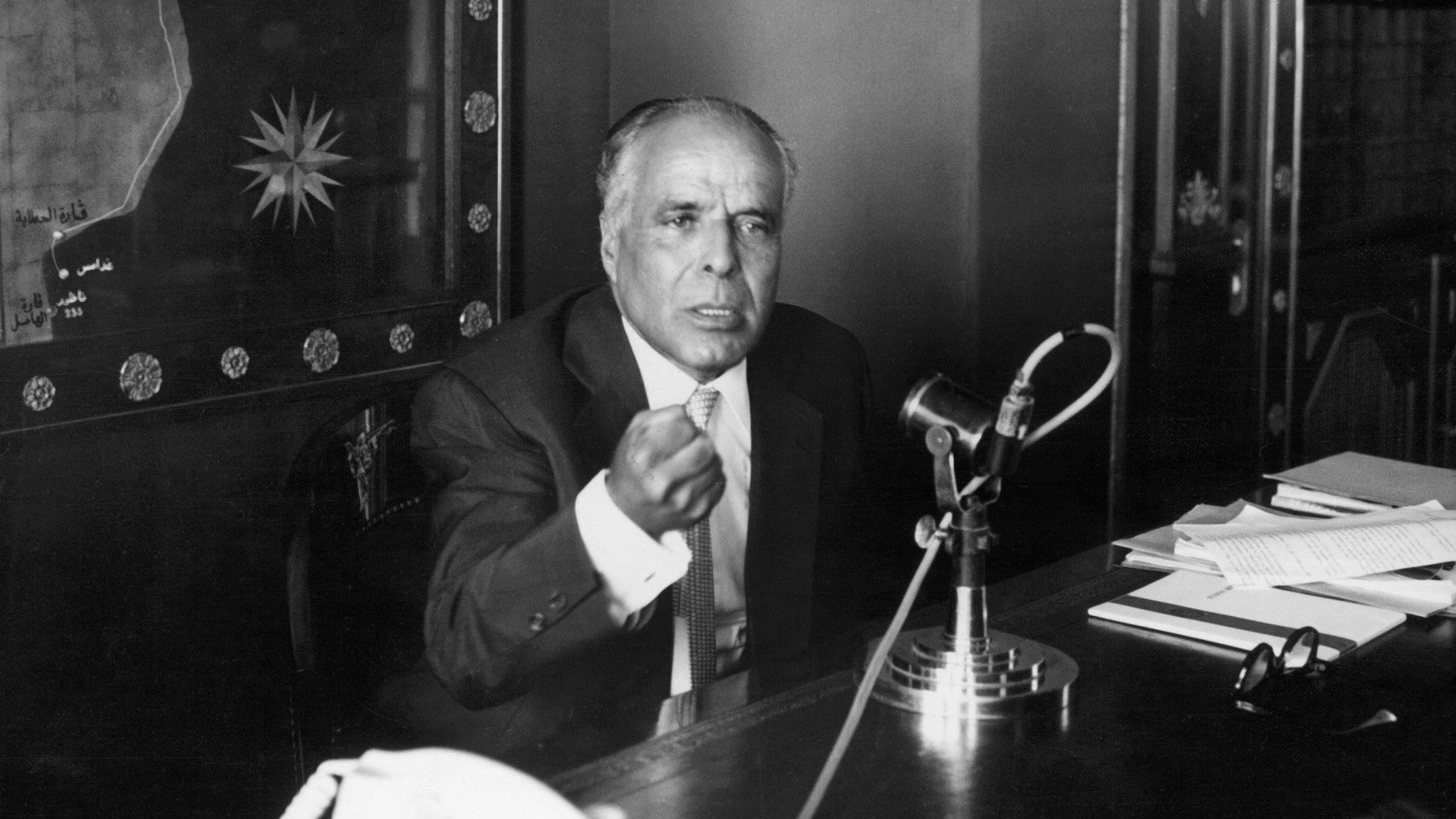 Homme en costume, gesticulant, devant un microphone et un bureau rempli de documents.