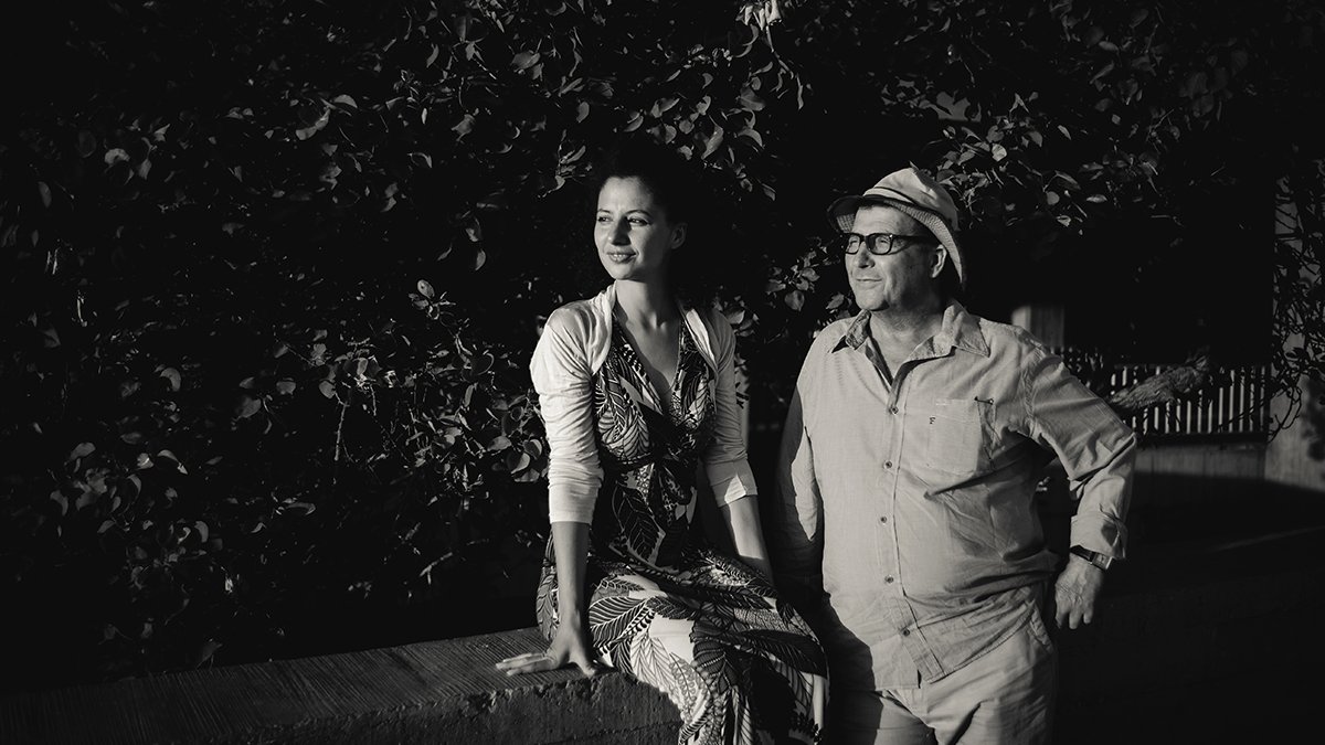 L'image montre une femme et un homme, tous deux dans un cadre extérieur, probablement près d'une plante ou d'un mur recouvert de verdure. La femme, habillée d'une robe imprimée, se tient assise sur un muret avec une posture détendue et un regard pensif. L'homme, vêtu d'un chemisier léger et d'un chapeau, se tient à côté d'elle avec un sourire, ajoutant à l'atmosphère conviviale de la scène. Le contraste entre les ombres et la lumière crée une ambiance artistique et intemporelle, en noir et blanc.