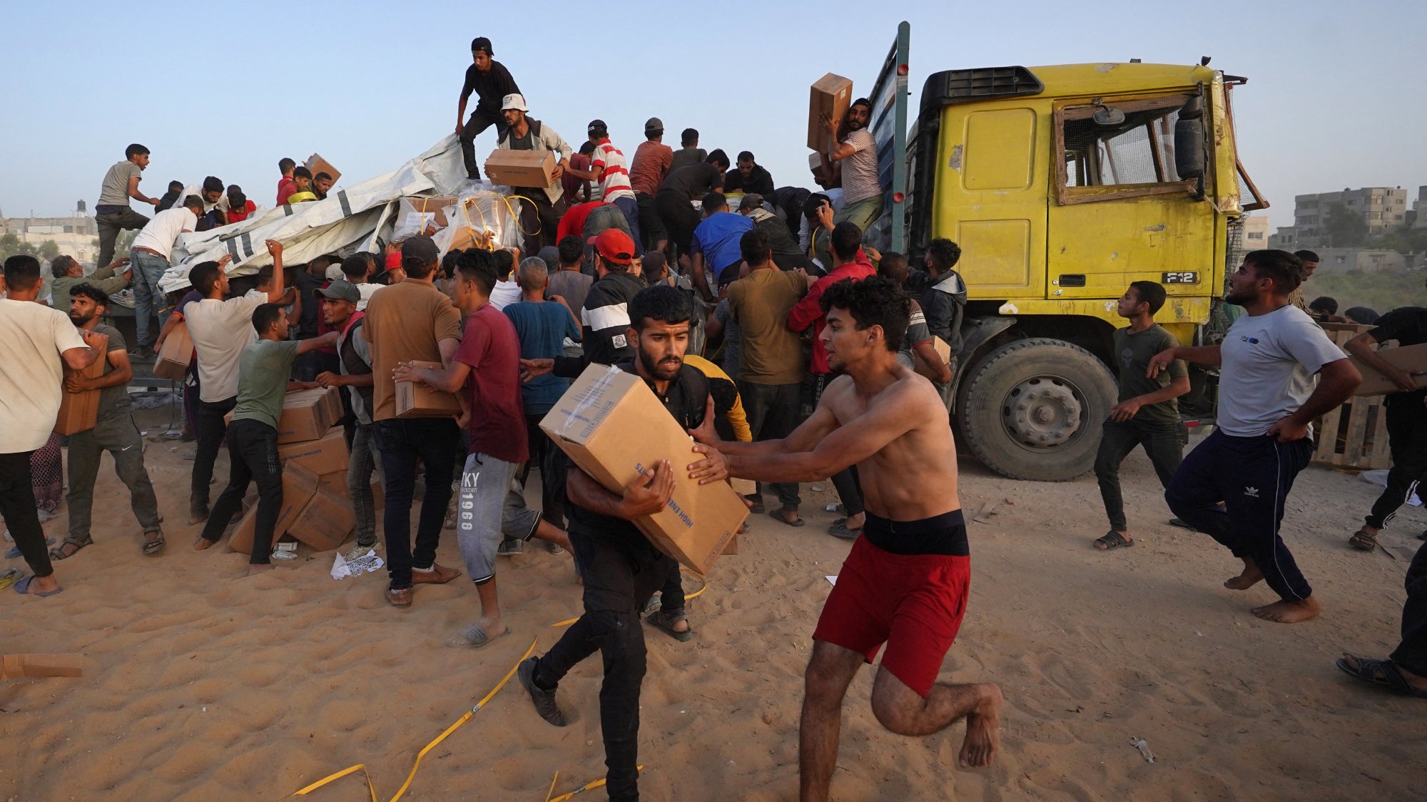L'image montre une scène animée où de nombreuses personnes semblent s'affairer autour d'un camion jaune. Des individus portent des boîtes et se déplacent rapidement, certains courant, tandis que d'autres semblent aider à décharger le camion. L'environnement est désertique, avec du sable au sol. L'atmosphère est chargée d'activité et d'urgence, indiquant qu'ils sont probablement en train de distribuer de l'aide ou des provisions.