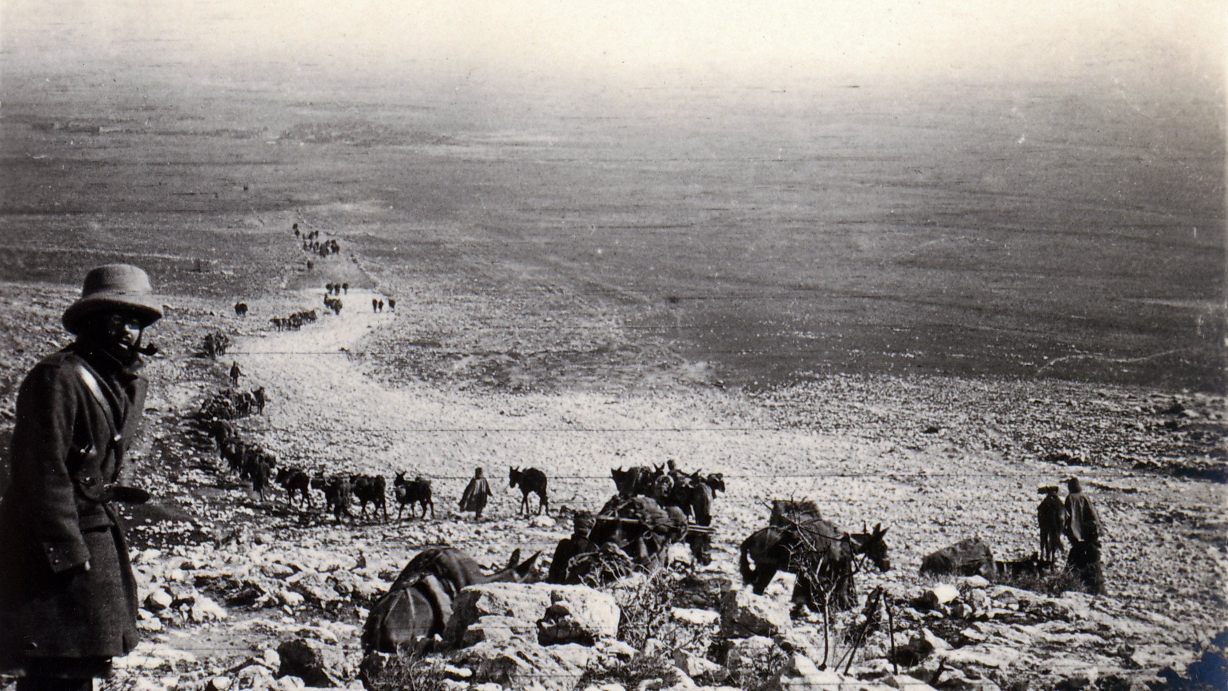 L'image montre un paysage désertique avec un groupe de personnes et d'animaux, probablement des mulets ou des ânes, qui avancent sur un chemin rocailleux. À l'avant, un homme en manteau et en chapeau observe la scène. Au loin, on peut voir une longue file de silhouette se déplaçant à travers des terrains arides. L'atmosphère est presque nostalgique, évoquant un temps passé.