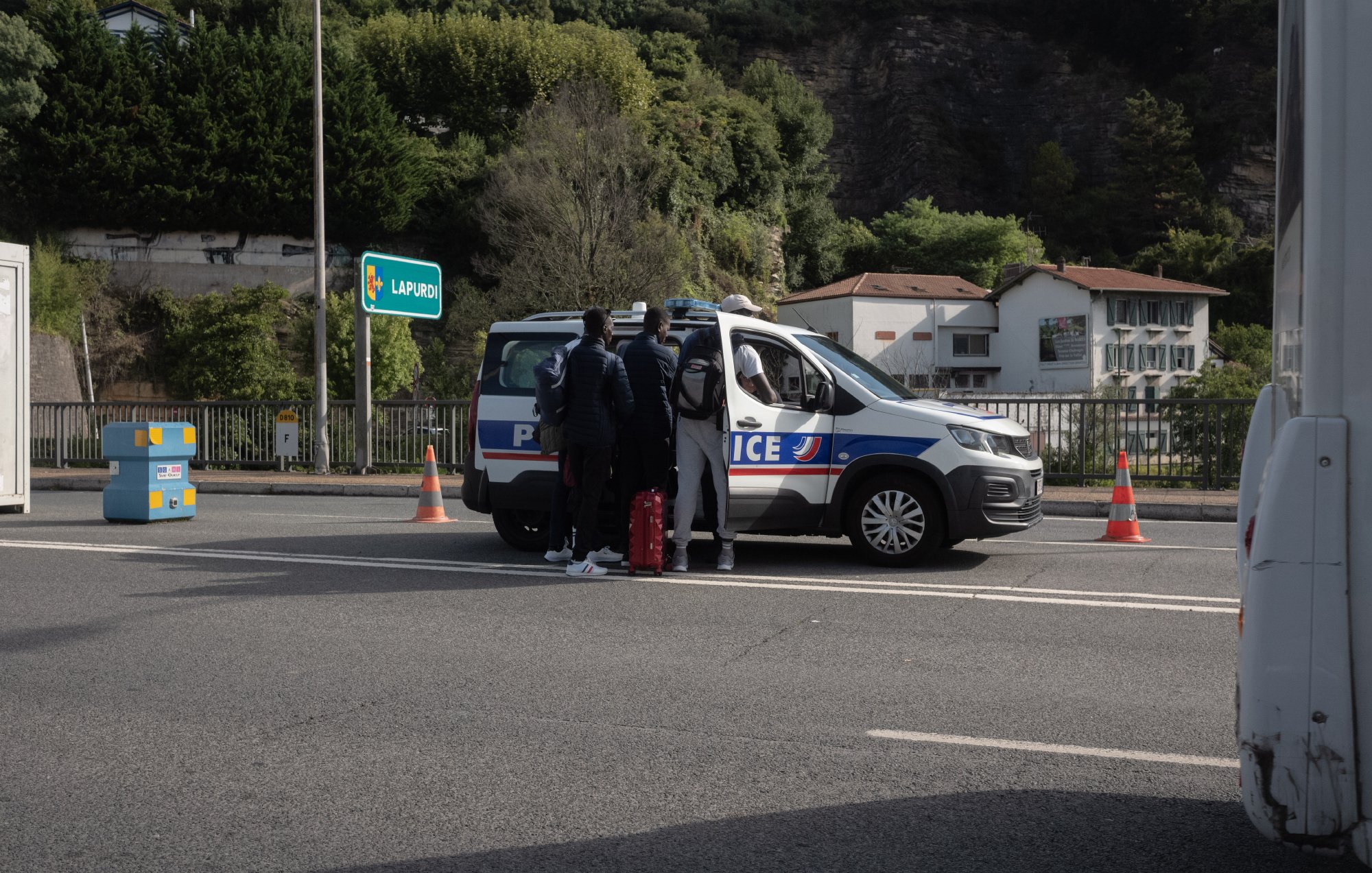 L'image montre une scène sur une route, où un véhicule de police est stationné. Des personnes se tiennent près de la voiture de police, probablement en train de discuter avec les agents. En arrière-plan, on peut voir quelques bâtiments et de la végétation. Des cônes de signalisation sont visibles sur la voie, indiquant peut-être une opération de contrôle ou de sécurité.