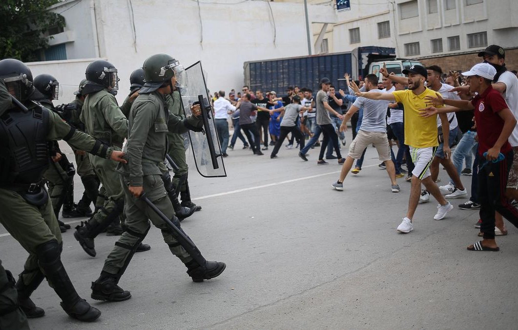 L'image montre une scène de confrontation entre des agents de sécurité en uniforme, portant des casques et des protections, et un groupe de manifestants. Les policiers avancent avec des boucliers, tandis que des personnes dans le public semblent manifester avec des gestes de défi. L'atmosphère est tendue, et le cadre urbain suggère un rassemblement intense, possiblement dans un contexte de protestation ou de manifestation. Les participants sont habillés de manière casual, et on peut voir une diversité dans leurs attitudes et leurs réactions face à la situation.