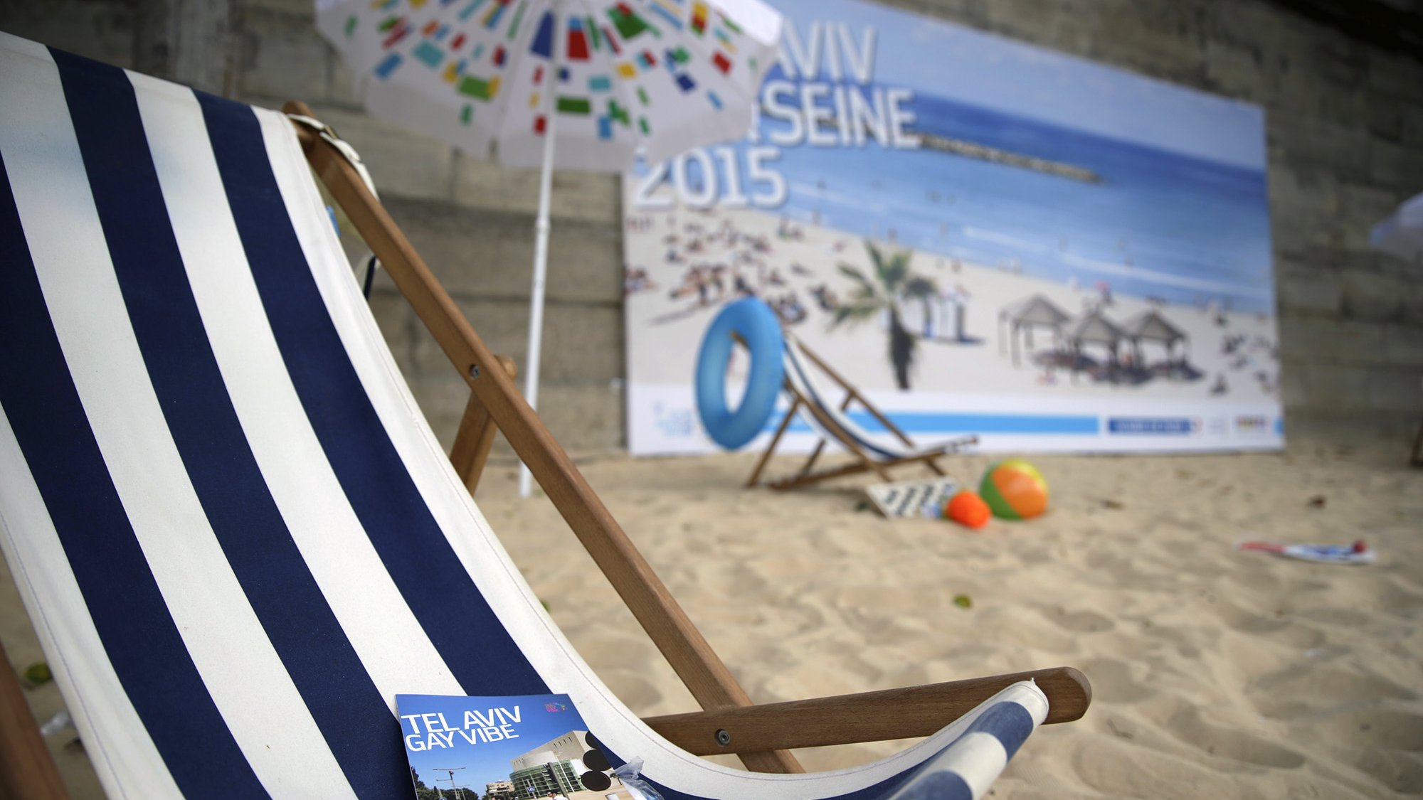 L'image montre une scène de plage avec un transat en bois rayé. Il y a des brochures posées sur le transat, portant des titres promouvant Tel Aviv et ses attractions. En arrière-plan, on peut voir un grand panneau affichant une ambiance estivale, suggérant une plage animée. Le sol est recouvert de sable, et on distingue également des objets colorés comme des ballons. Des parasols, ainsi qu'une vue sur la mer, ajoutent une atmosphère estivale à l'ensemble.