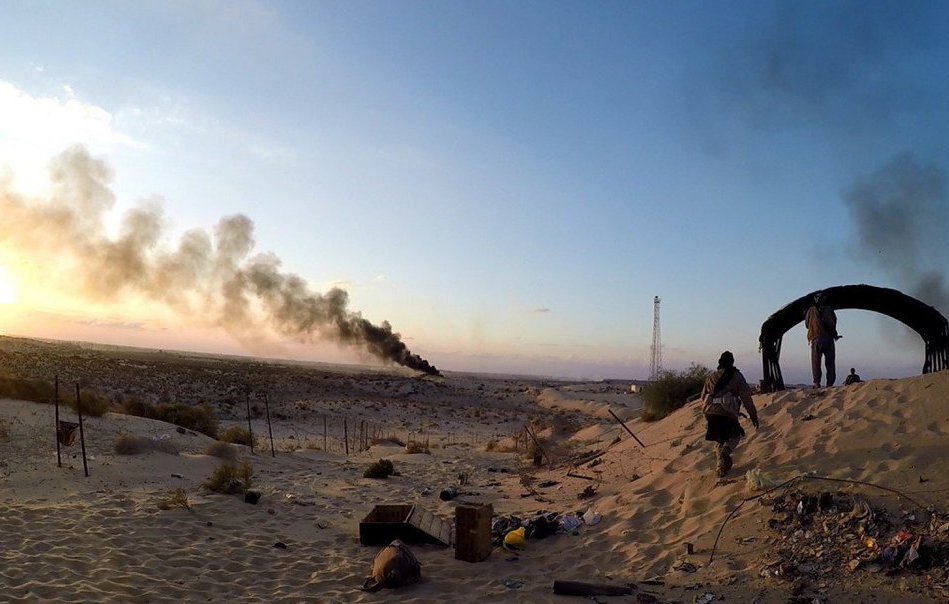 L'image montre un paysage désertique avec une ambiance dramatique. À l'arrière-plan, on peut voir des colonnes de fumée noire s'élevant dans le ciel, suggérant qu'il y a un incendie ou une explosion. Au premier plan, des silhouettes de personnes évoluent dans le sable, probablement en train d'explorer ou de surveiller la zone. La lumière dorée du coucher de soleil crée un contraste frappant avec l'obscurité de la fumée, ajoutant à l'atmosphère générale de tension et de désolation. Le décor semble abandonné, avec quelques débris éparpillés sur le sol.
