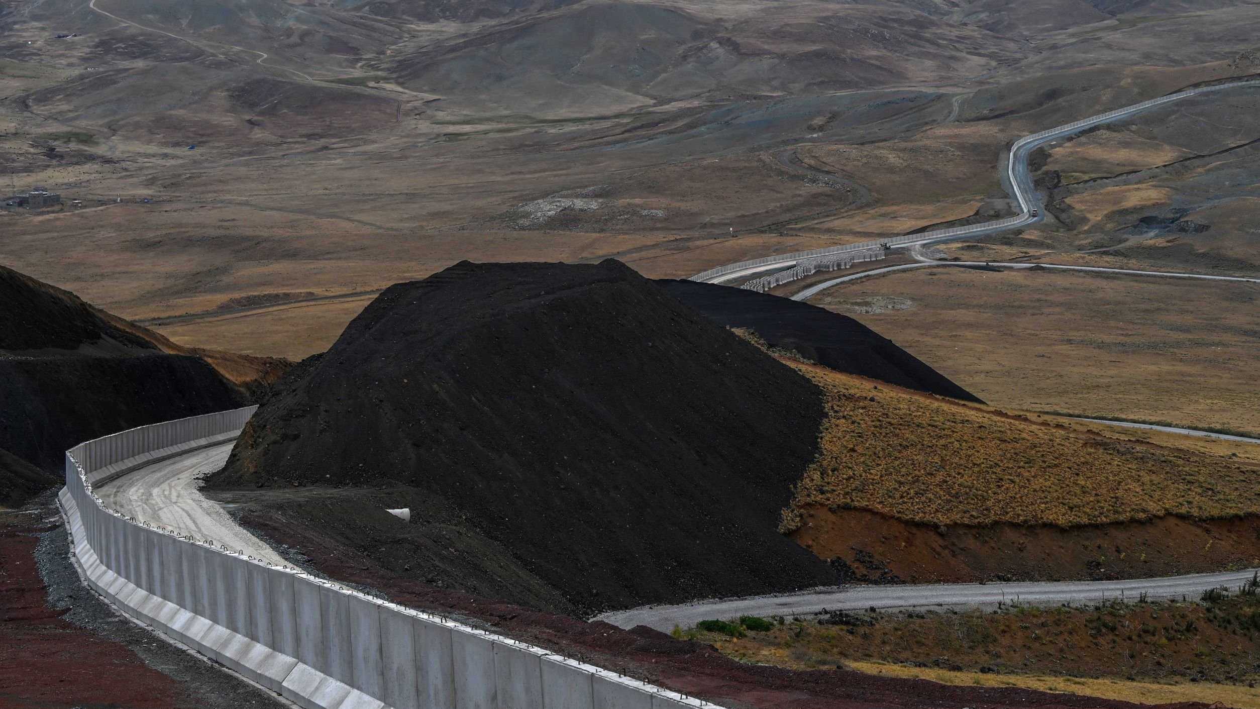 L'image montre un paysage vaste et aride, avec des collines et des montagnes en arrière-plan. Au premier plan, il y a un mur de béton qui serpente le long du sol, suivi d'une route qui s'étend dans la vallée. Le terrain est principalement composé de terre brune et de végétation clairsemée, avec des déclivités et des pentes. L'ambiance générale semble désertique et isolée.